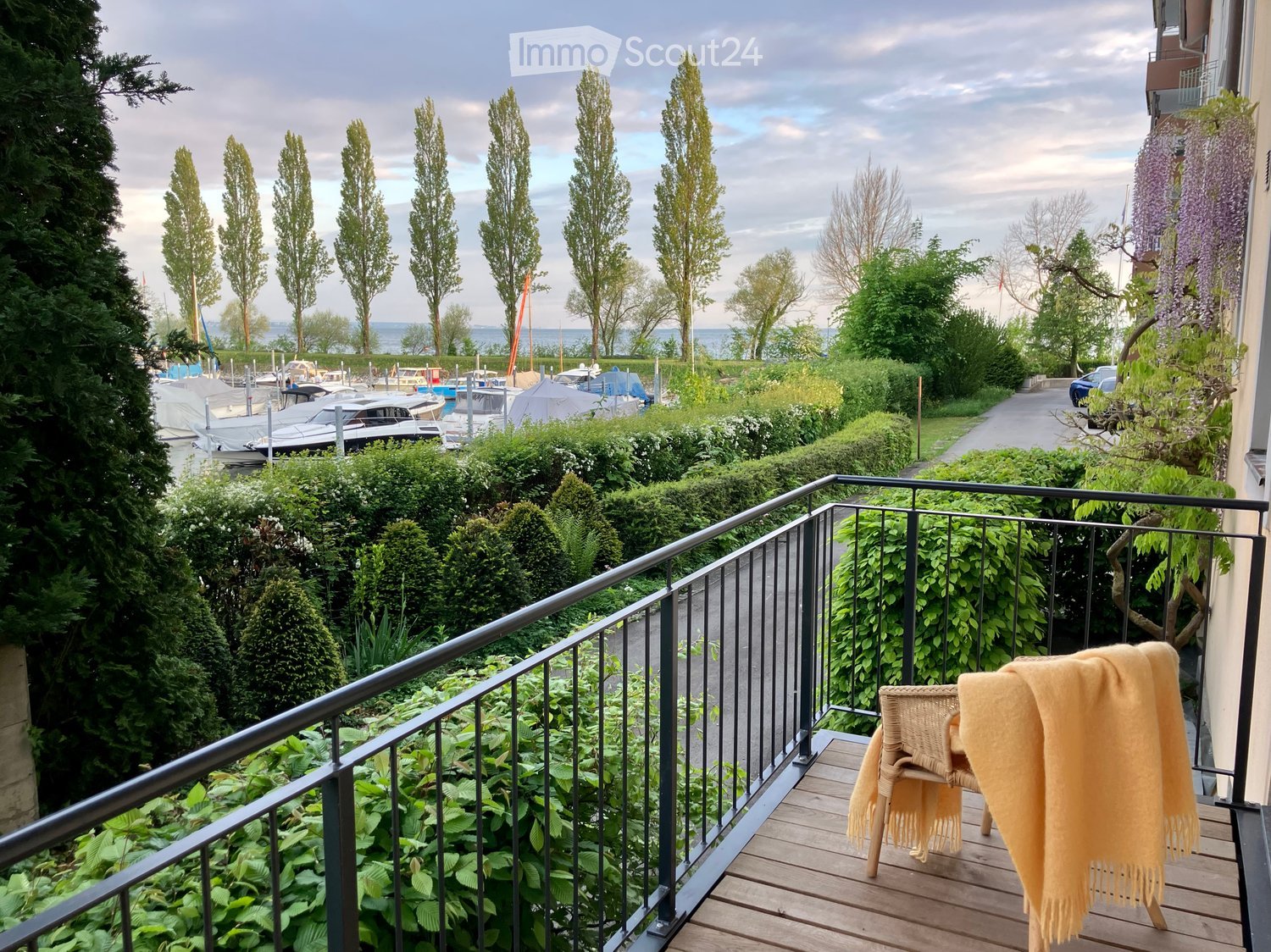 Balcony with wooden decking, metal railing, and a view of a harbor with boats and trees.