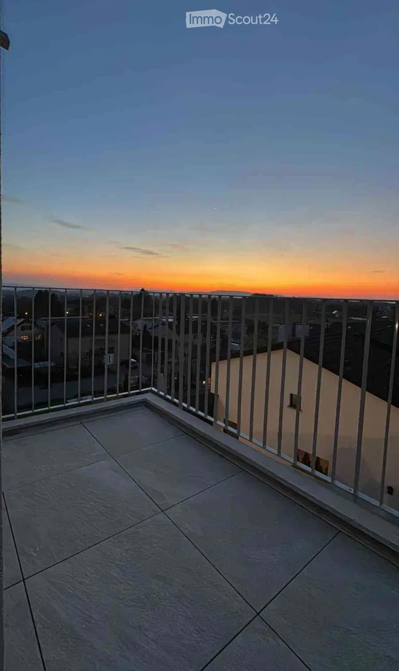 Balcony with a modern metal railing, tiled floor, and a view of the cityscape at sunset.