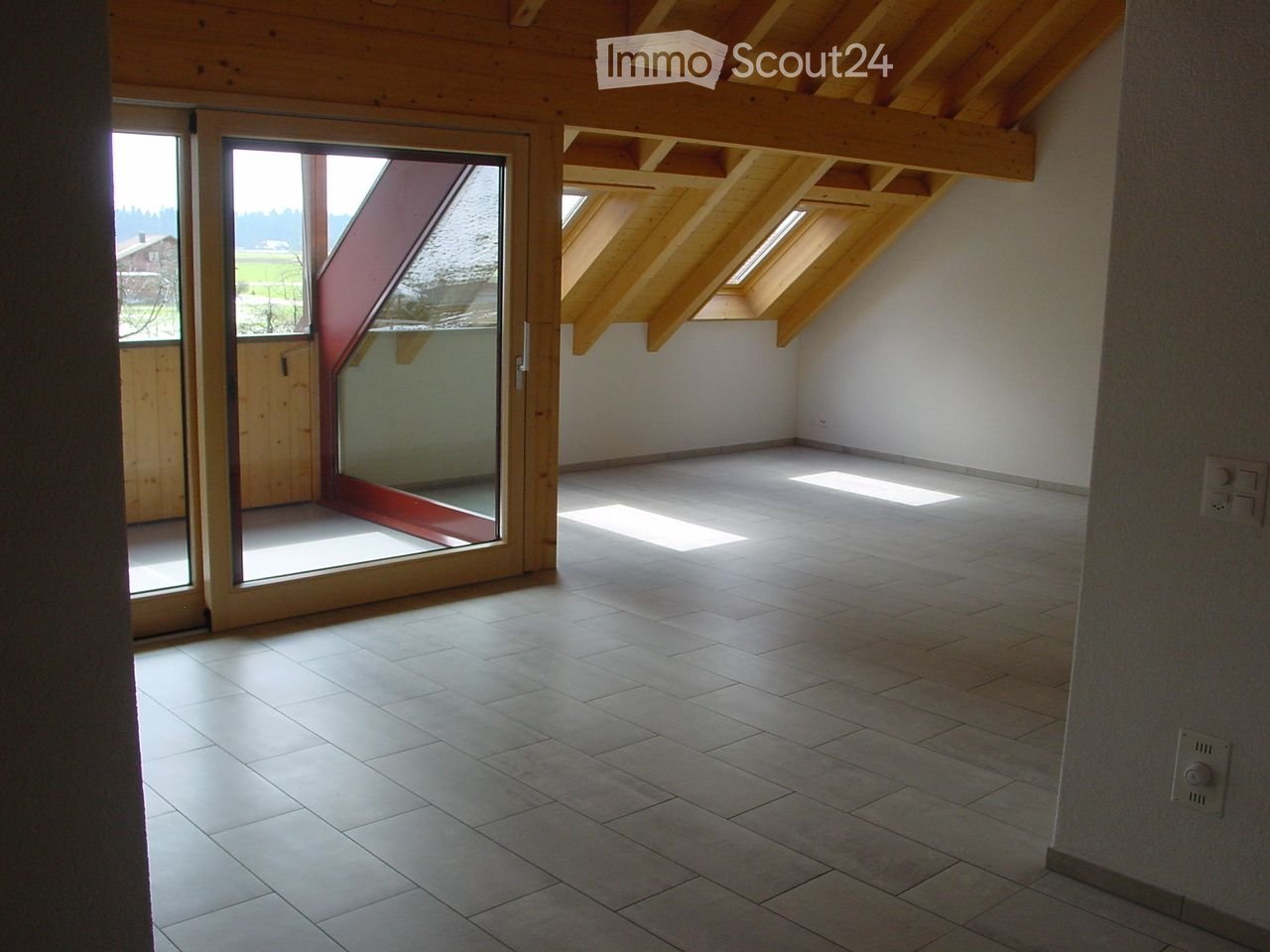 Empty room with exposed wooden beams, tiled floor, large sliding glass door leading to a balcony, and skylights allowing natural light.