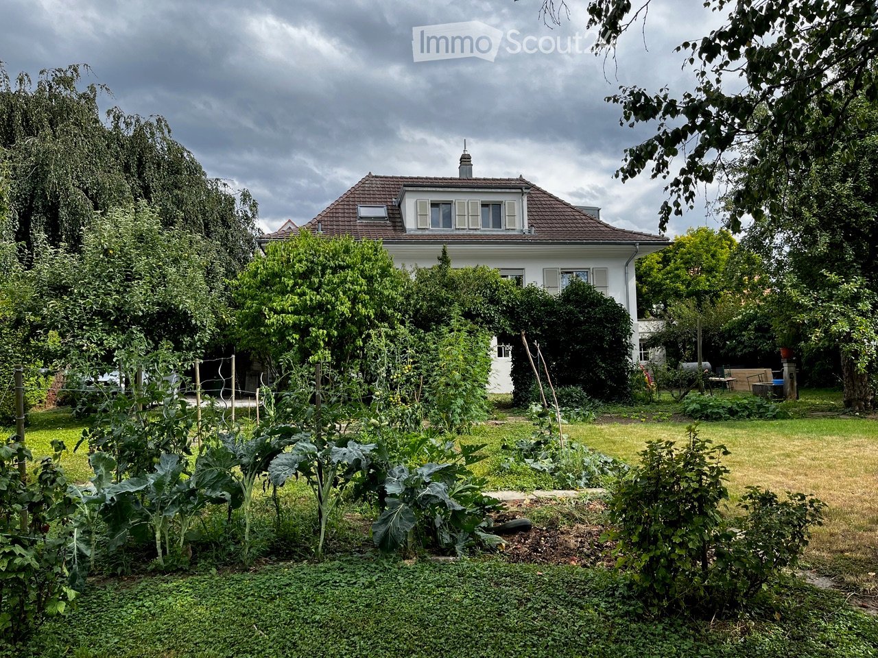 A well-maintained garden with a variety of plants and vegetables, featuring a white house with a red-tiled roof in the background.