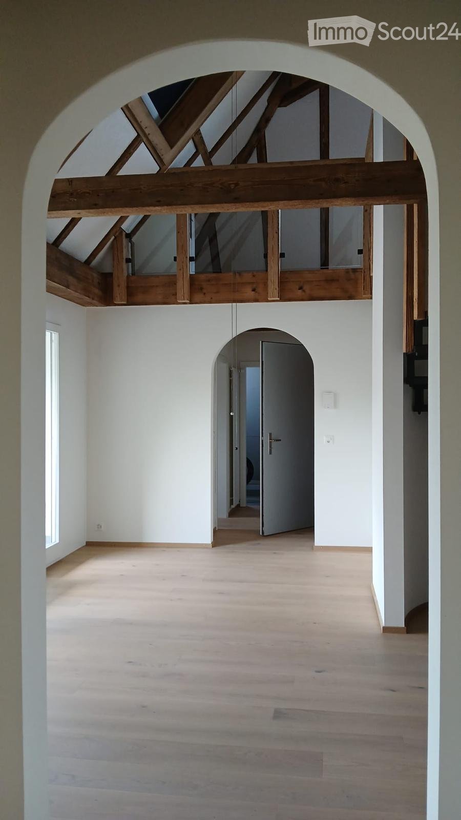 Empty room with wooden flooring, white walls, and exposed wooden beams in the ceiling. A doorway leads to another room.