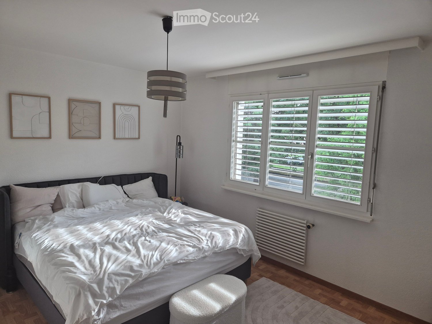 Bright bedroom with a large bed, white bedding, a small stool, radiator, and three picture frames on the wall. Large windows with blinds let in natural light.