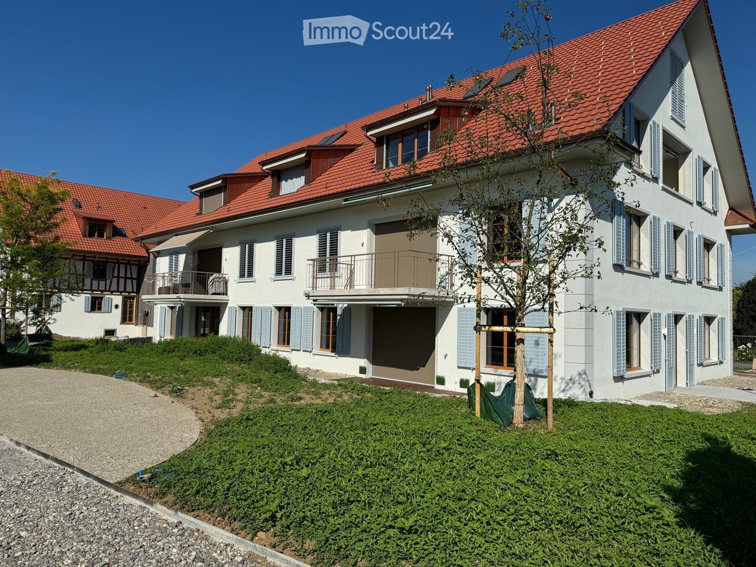 Two-story house with red-tiled roof, white walls, multiple windows with shutters, and a balcony on the upper floor.