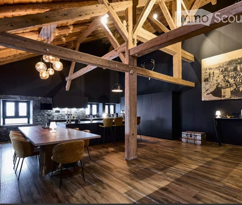 Open-concept dining and kitchen area with exposed wooden beams, wooden flooring, and modern lighting. Features a large wooden dining table with chairs, a kitchen island with stools, pendant lights, and a large picture frame on the wall.