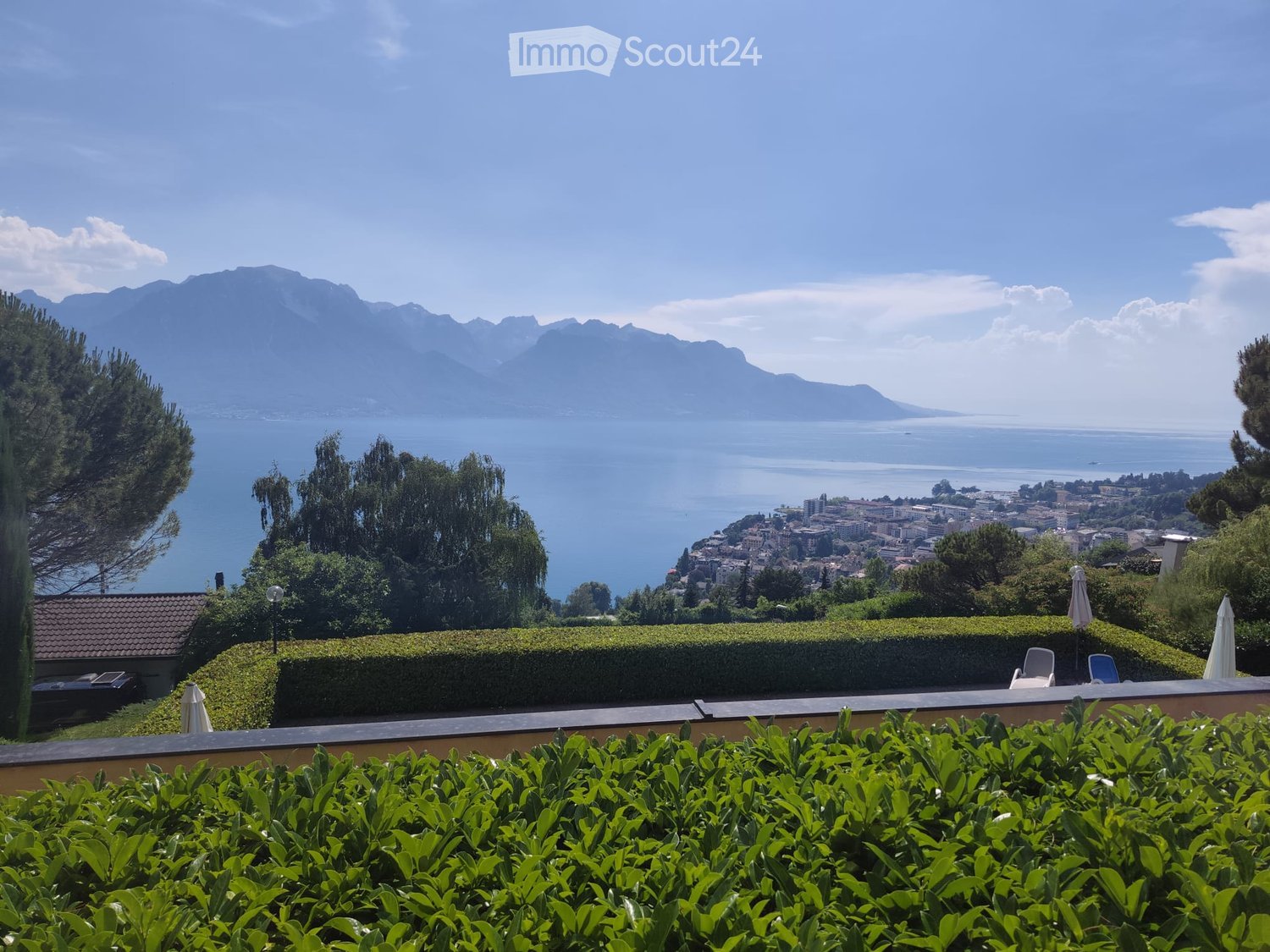 A scenic garden terrace overlooking a lake and distant mountains, with neatly trimmed hedges, outdoor chairs, and umbrellas.