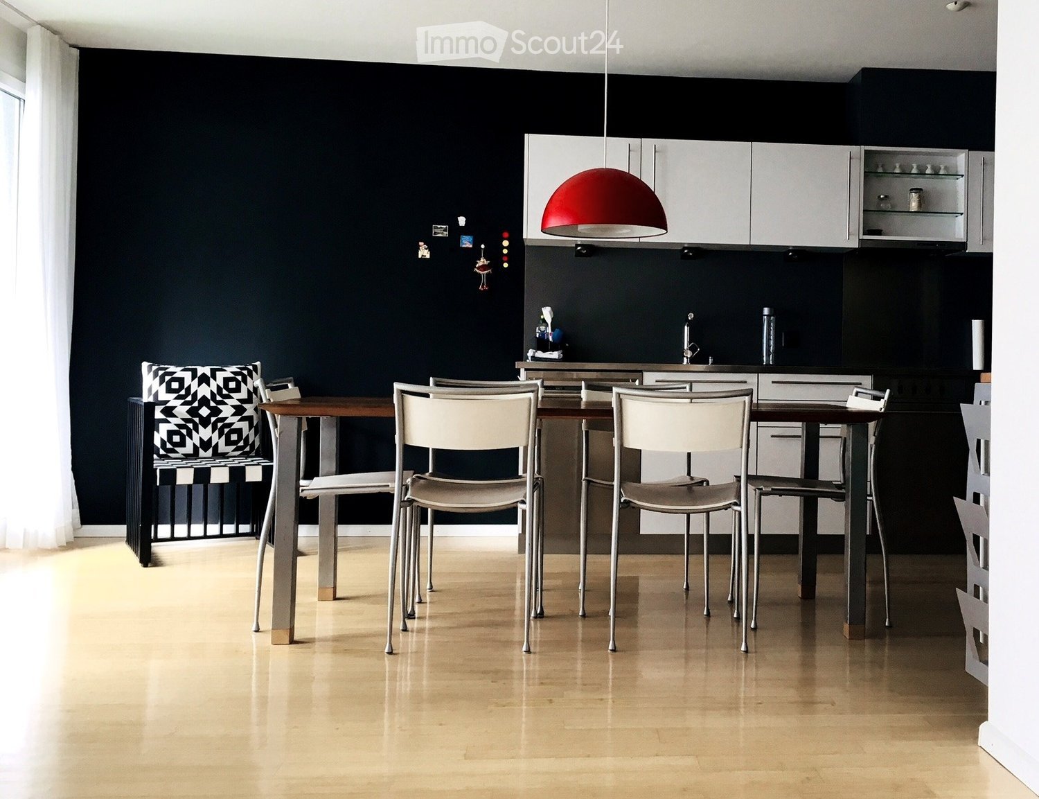 Modern dining area with a wooden table, white chairs, and a sleek kitchen area featuring white cabinets and a red pendant light.