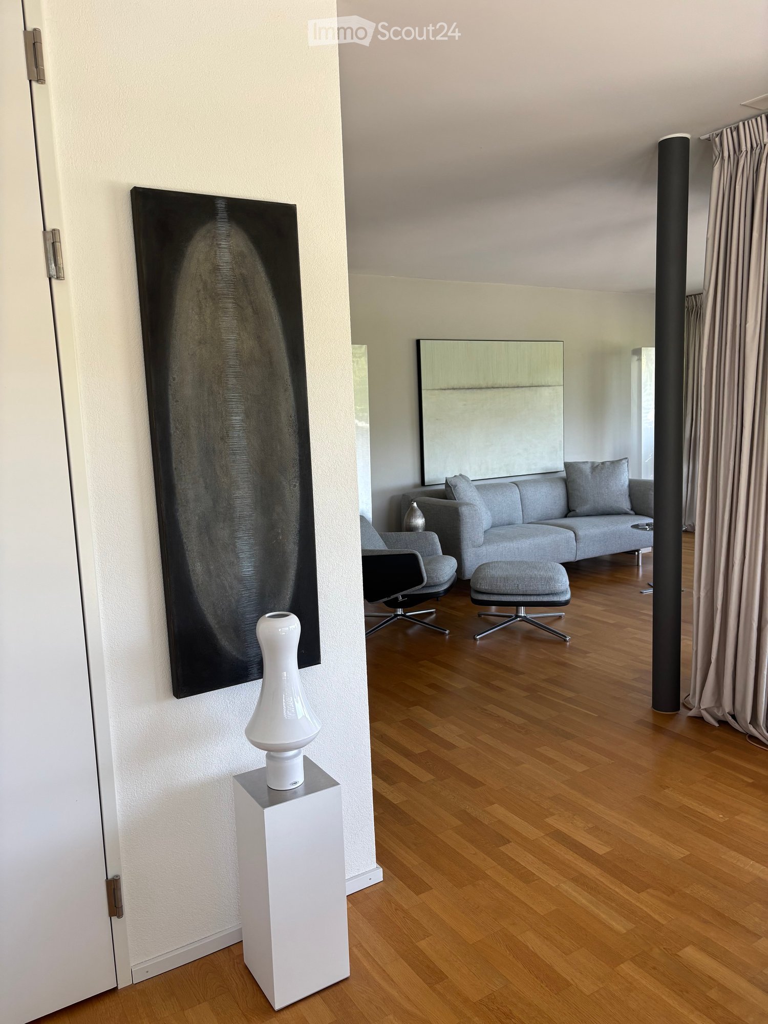 Modern living room with hardwood flooring, grey sofa, ottoman, chairs, a white vase on a table, and a large mirror on the wall.
