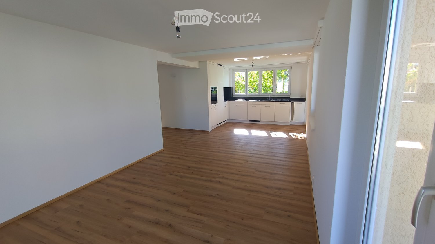 Empty room with wooden flooring and white walls, leading to a small kitchen area with white cabinets and black countertops.