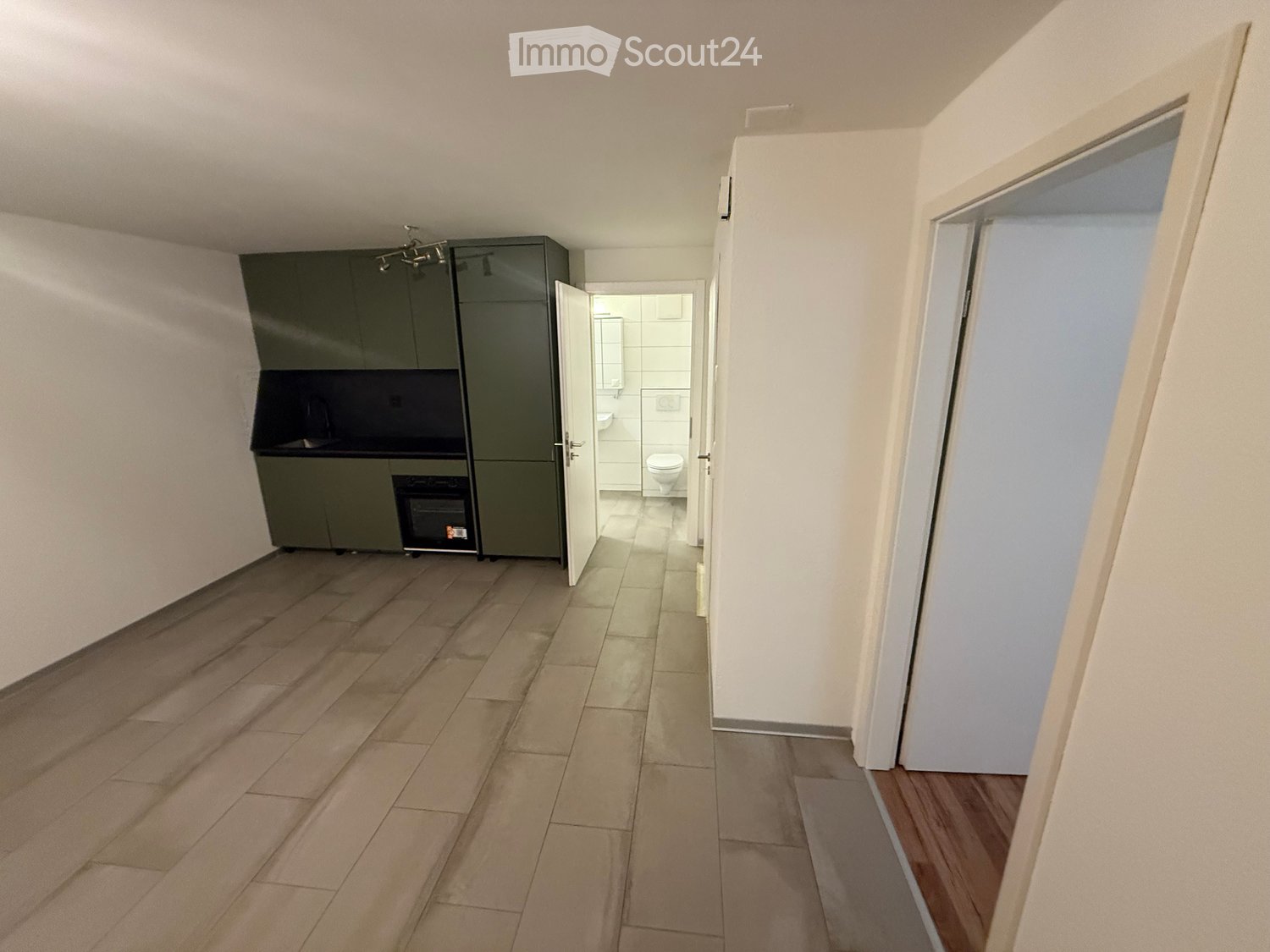 Empty hallway with tiled floor, white walls, and a small kitchen area with dark green cabinets and a sink, leading to a bathroom with a toilet and sink.