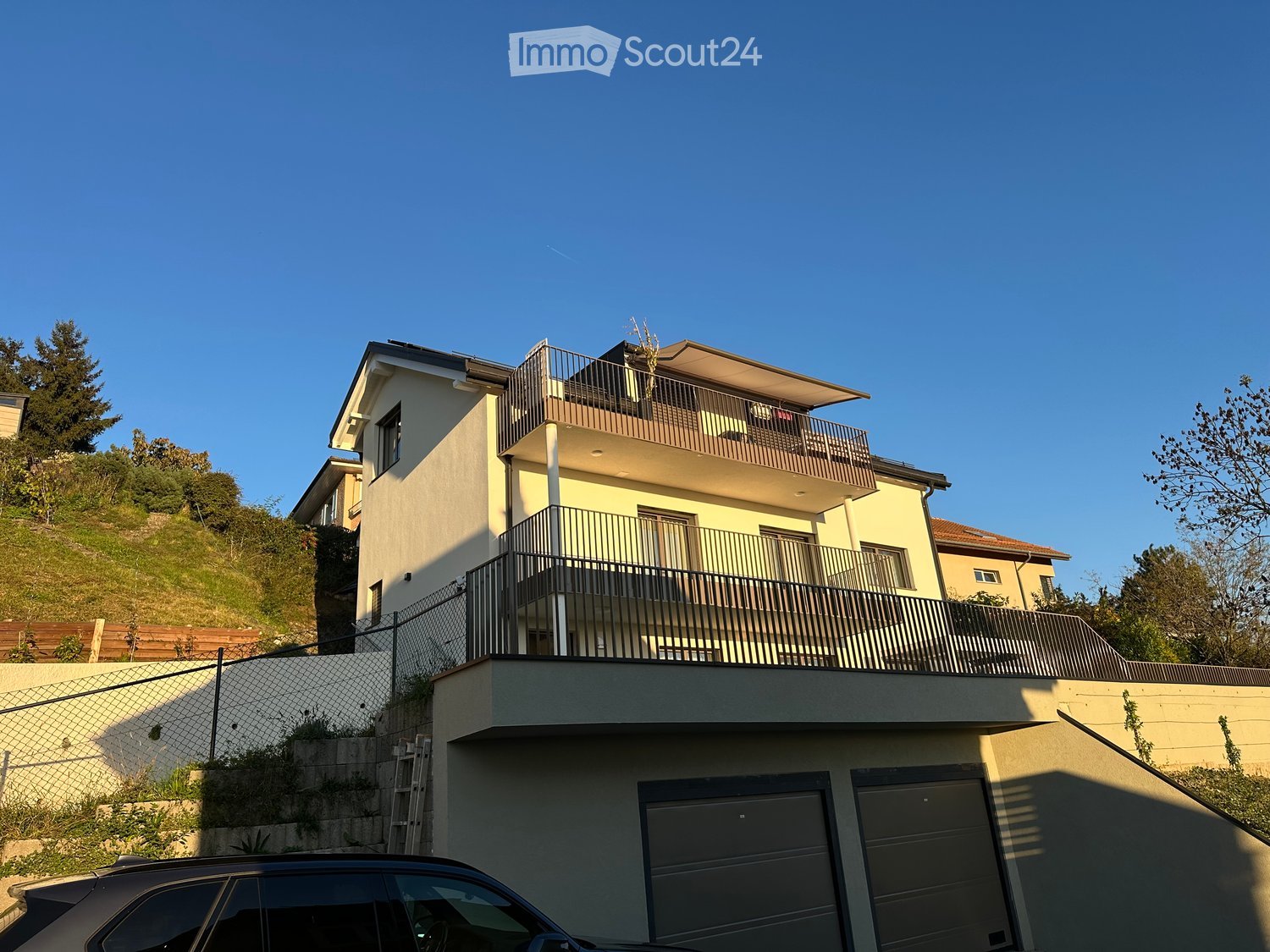 Modern two-story house with balconies, light yellow facade, and multiple windows. Features include a fenced yard, garage, and a raised ground floor.