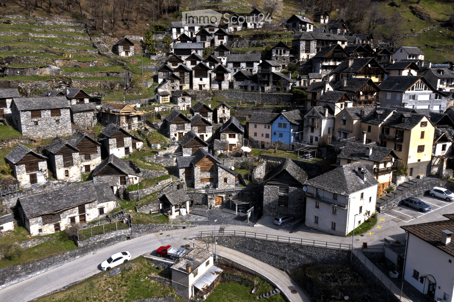 Aerial view of a small village with stone houses, narrow roads, and a few parked cars.