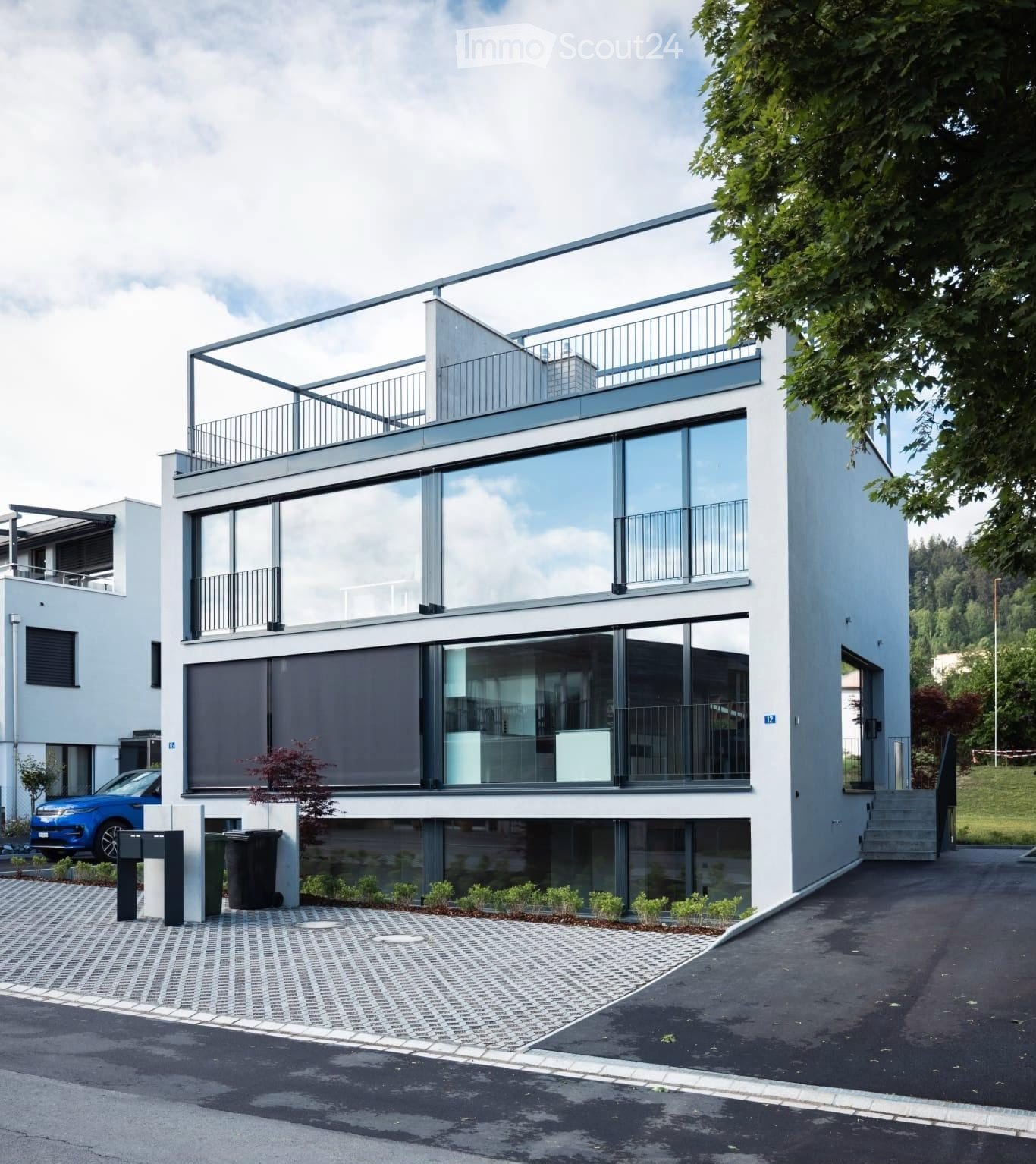 Modern two-story house with large glass windows, balconies, and a raised ground floor. Features a paved driveway, small garden area, and a blue car parked nearby.