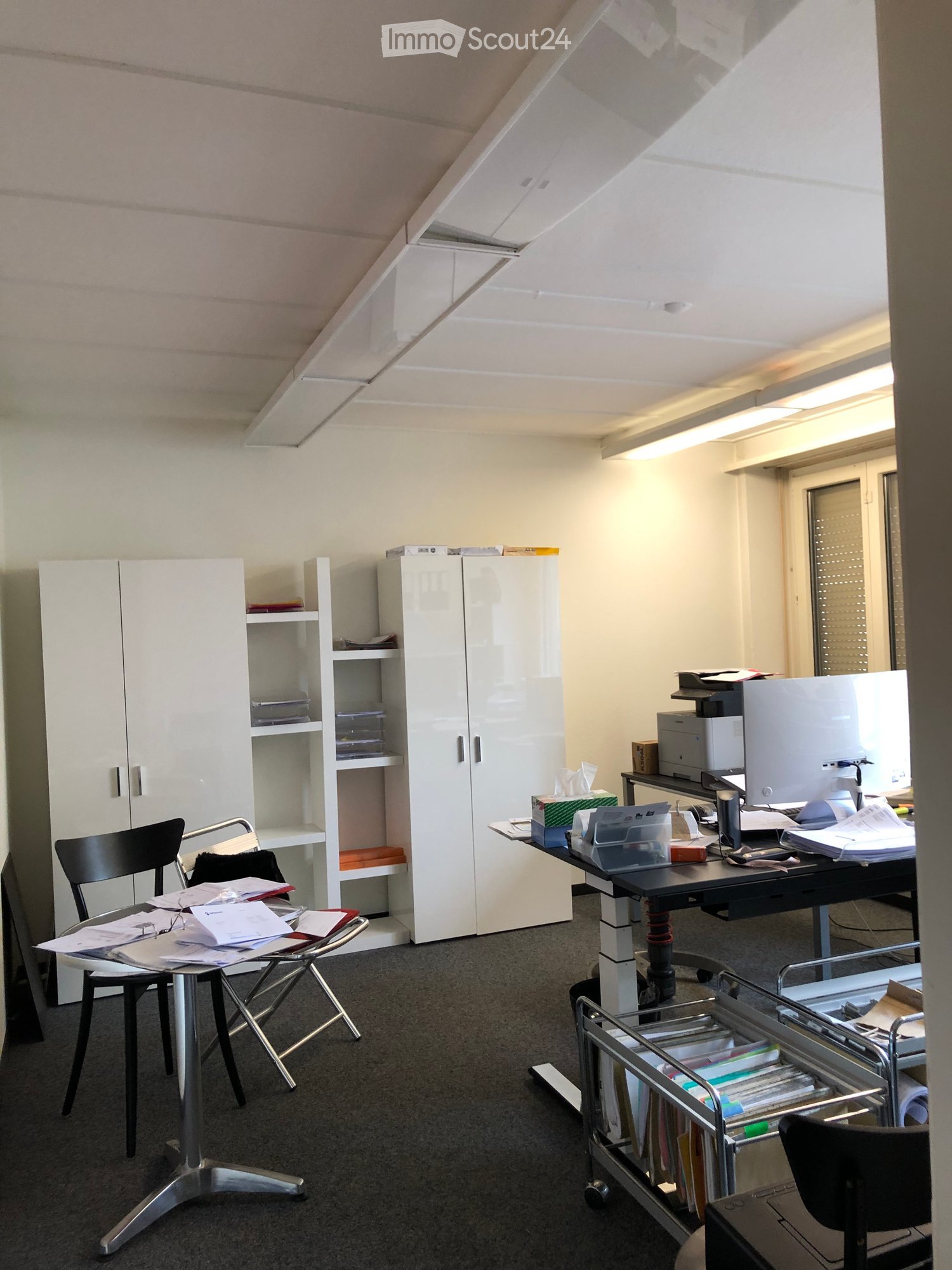 Office space with a slanted ceiling, white cabinets, shelves, a desk with a computer monitor, printer, and various office supplies, along with chairs and a rolling cart holding files.