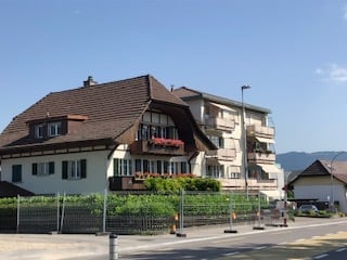 A multi-story residential building with a brown tiled roof, white walls, and multiple balconies. The building is surrounded by greenery and is situated on a street with a clear blue sky in the background.