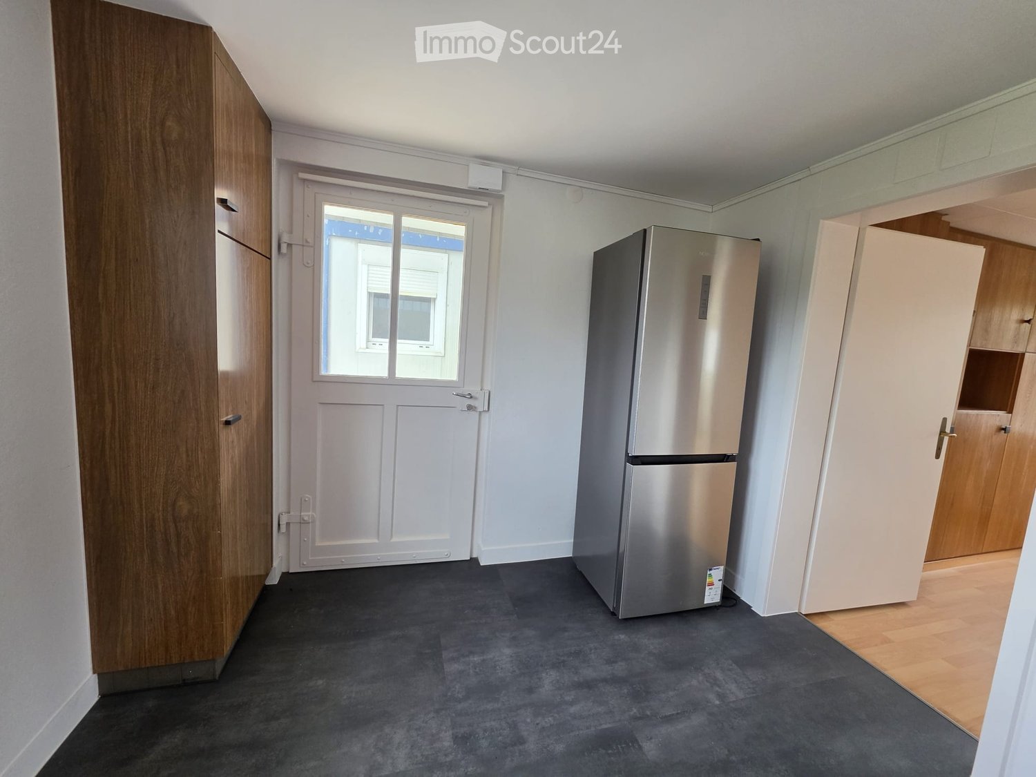 A small, furnished room with a stainless steel refrigerator, a wooden wardrobe, and a door leading outside.