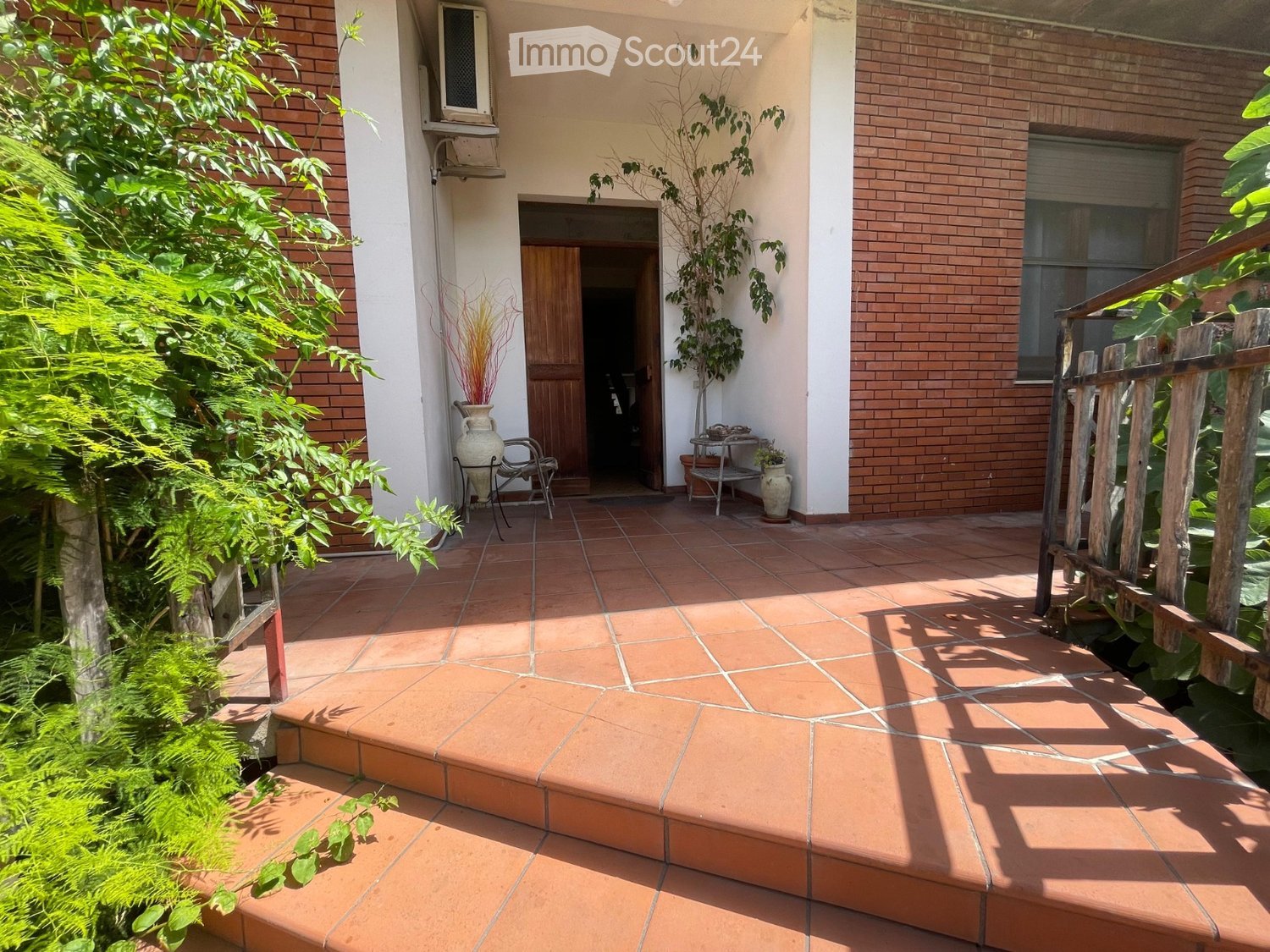 A tiled outdoor patio area with a small table, chairs, vases, and plants, leading to a wooden door entrance of a house.