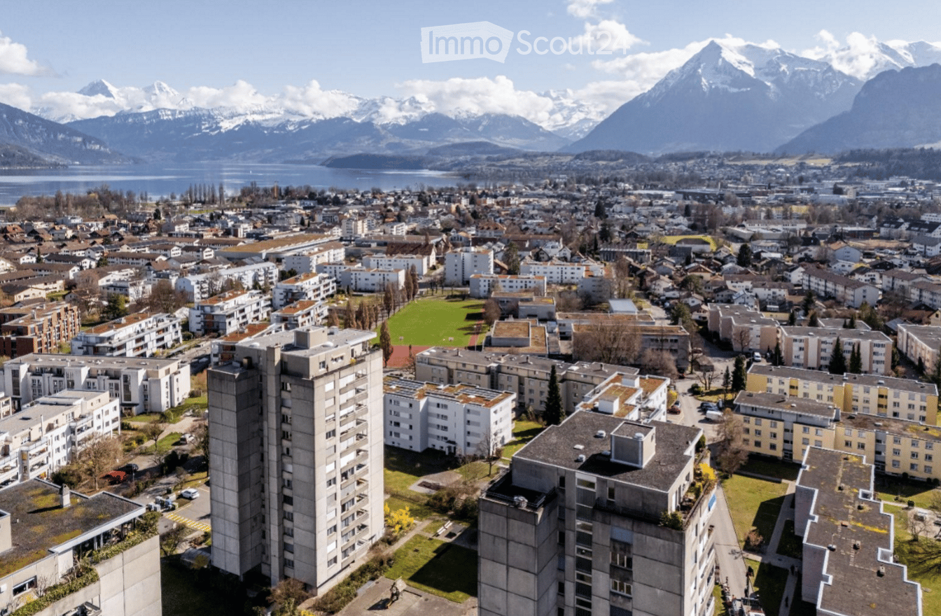 Aerial view of a residential area with apartment buildings, surrounded by green spaces and a scenic mountain backdrop.