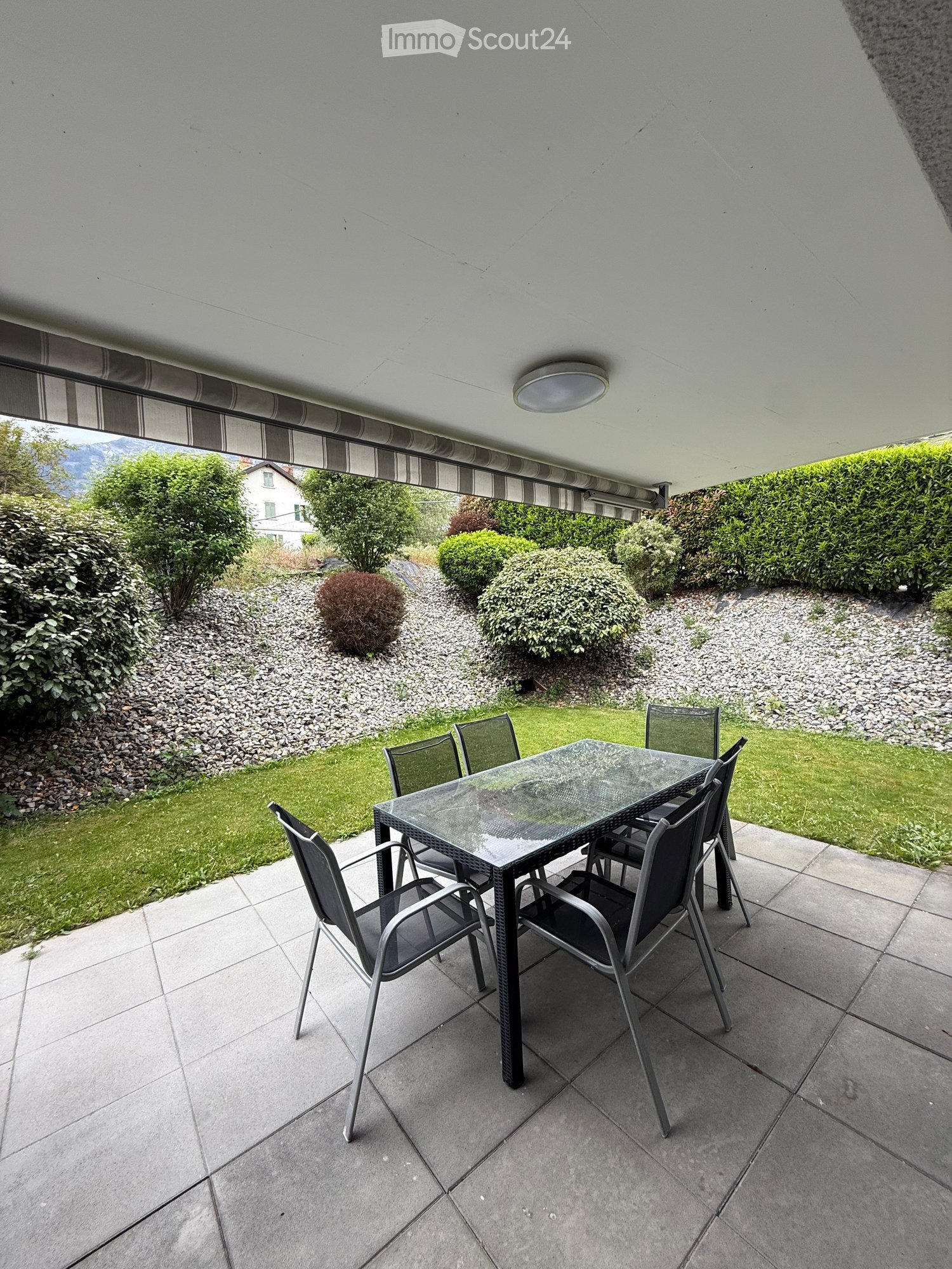 Balcony with a glass table and six chairs, surrounded by a well-maintained garden with shrubs and a gravel pathway.