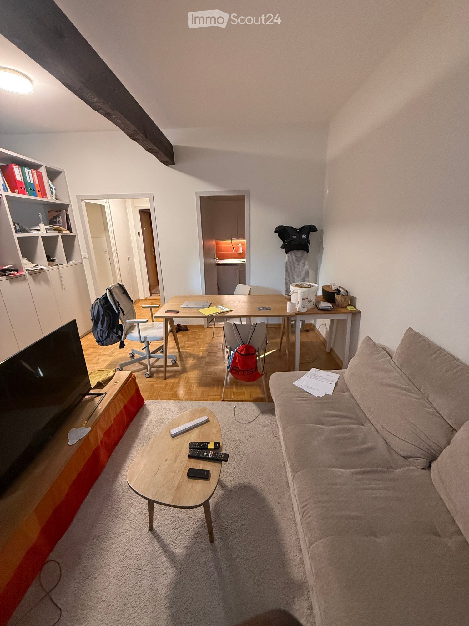A cozy living room with a sofa, coffee table, TV stand, and a small dining area with a table and chairs. Shelves with books and a backpack are visible, along with a kitchen area in the background.