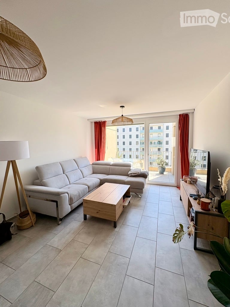 Modern living room with a grey sectional sofa, wooden coffee table, TV stand, floor lamp, and potted plants. Large windows with red curtains lead to a balcony.