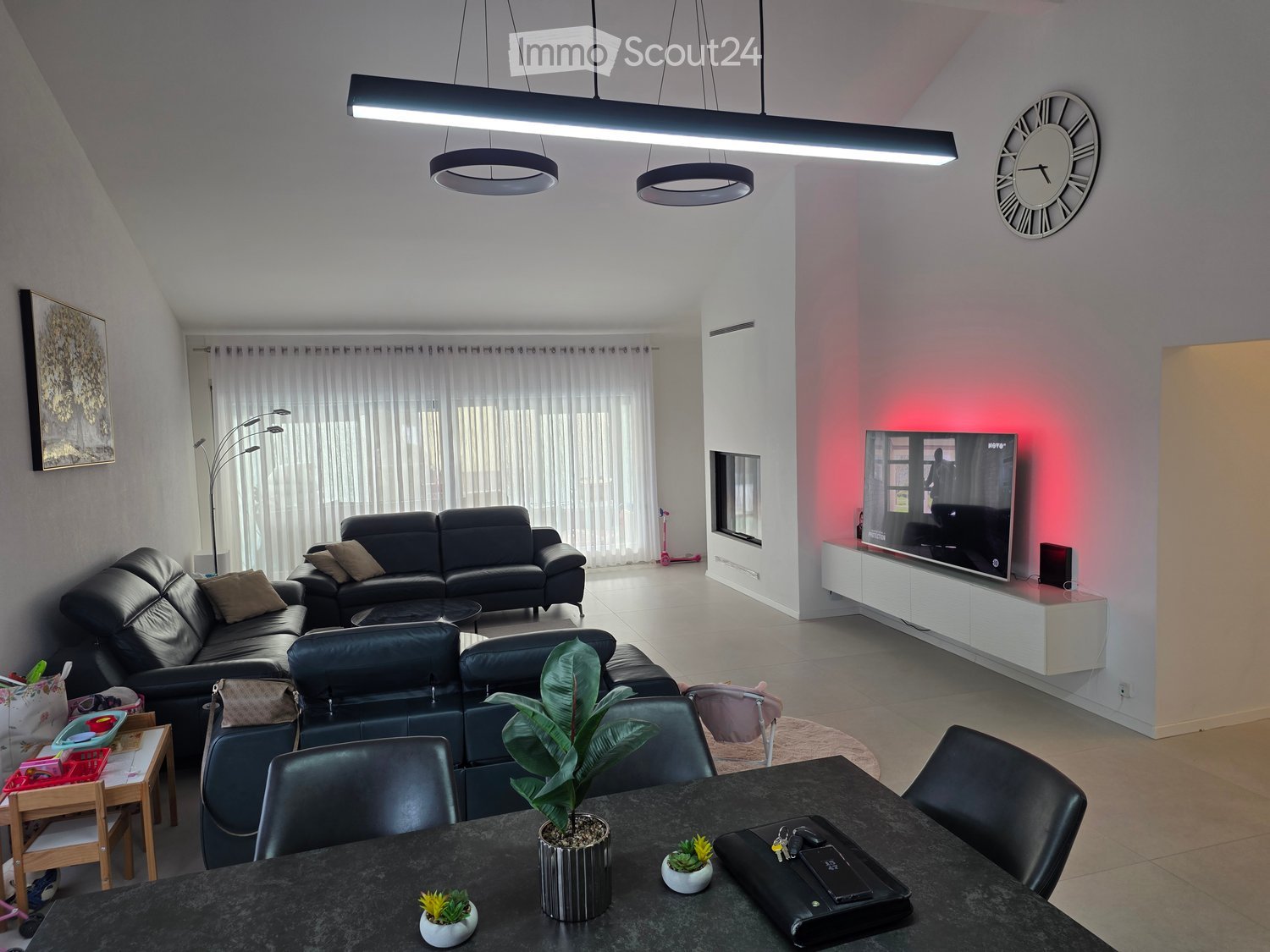 Modern living room with a black leather sofa, coffee table, dining table with chairs, TV, floor lamp, and decorative plants.