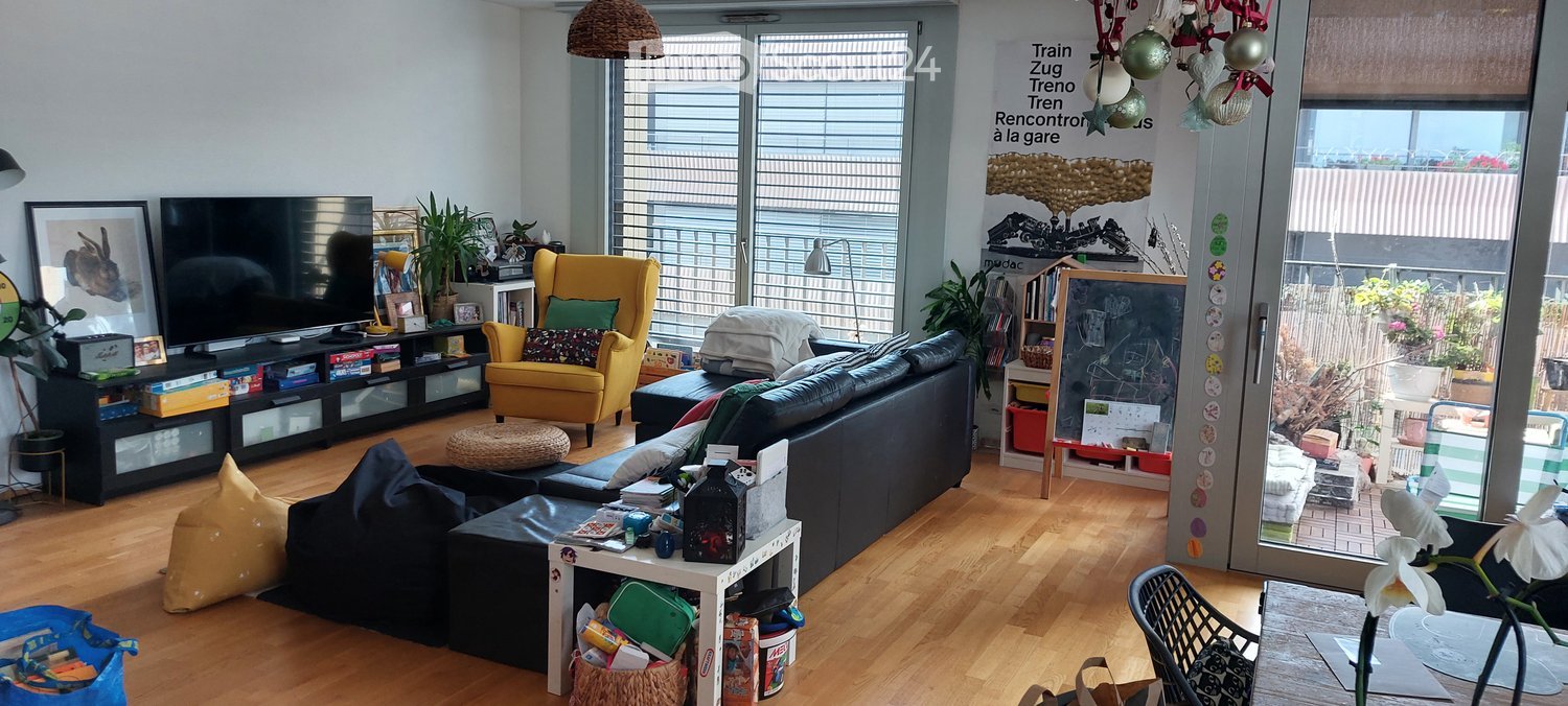 Modern living room with wooden flooring, large windows, a black sectional sofa, yellow armchair, coffee table, TV, and various decorative items including plants and artwork.