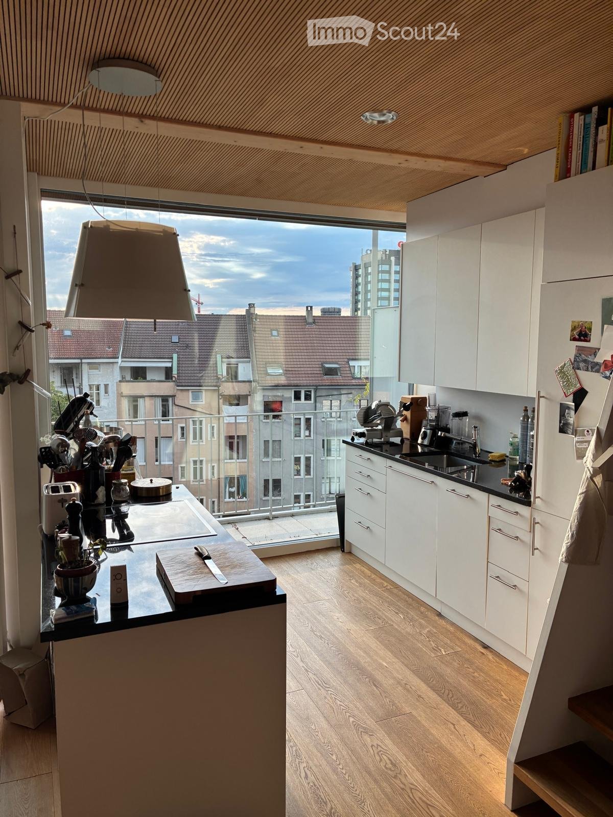 Modern kitchen with white cabinets, black countertops, wooden floor, and a large window overlooking a cityscape. Includes a sink, cutting board, knife, toaster, blender, and a refrigerator.
