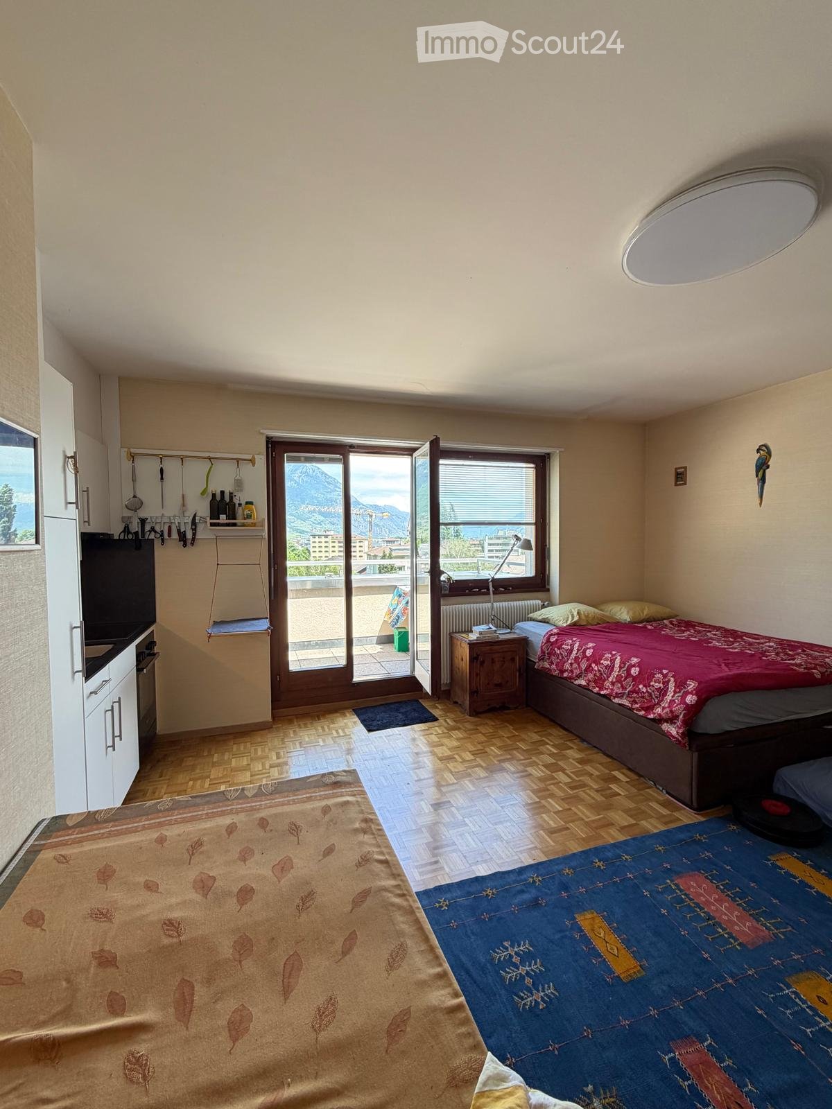 Bright bedroom with two beds, nightstand, lamp, and a small kitchen area with a fridge and screen. Large windows offer a scenic mountain view.