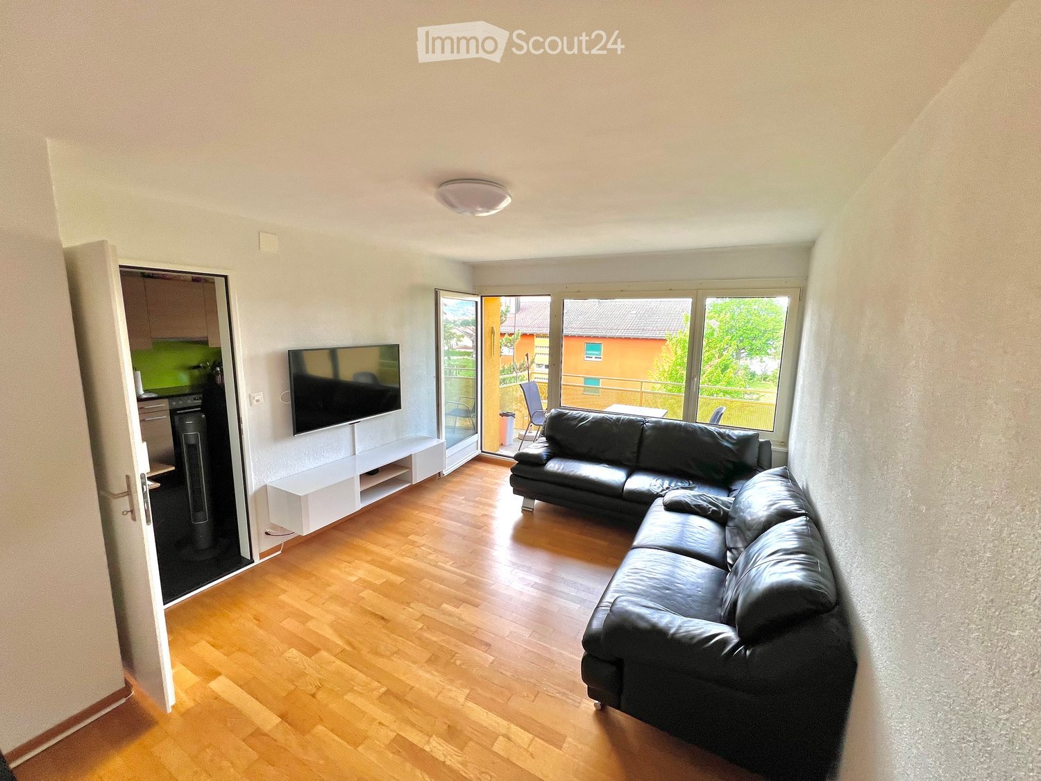 Modern living room with wooden flooring, a black leather sofa, a flat-screen TV on a white stand, and a sliding glass door leading to a balcony.
