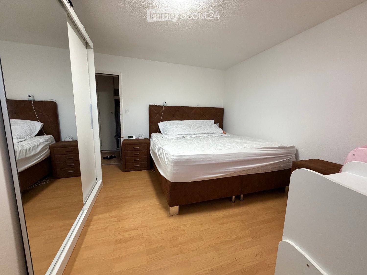 Cozy bedroom with two single beds, nightstands, a mirror, and a small chair, featuring light wooden flooring and white walls.