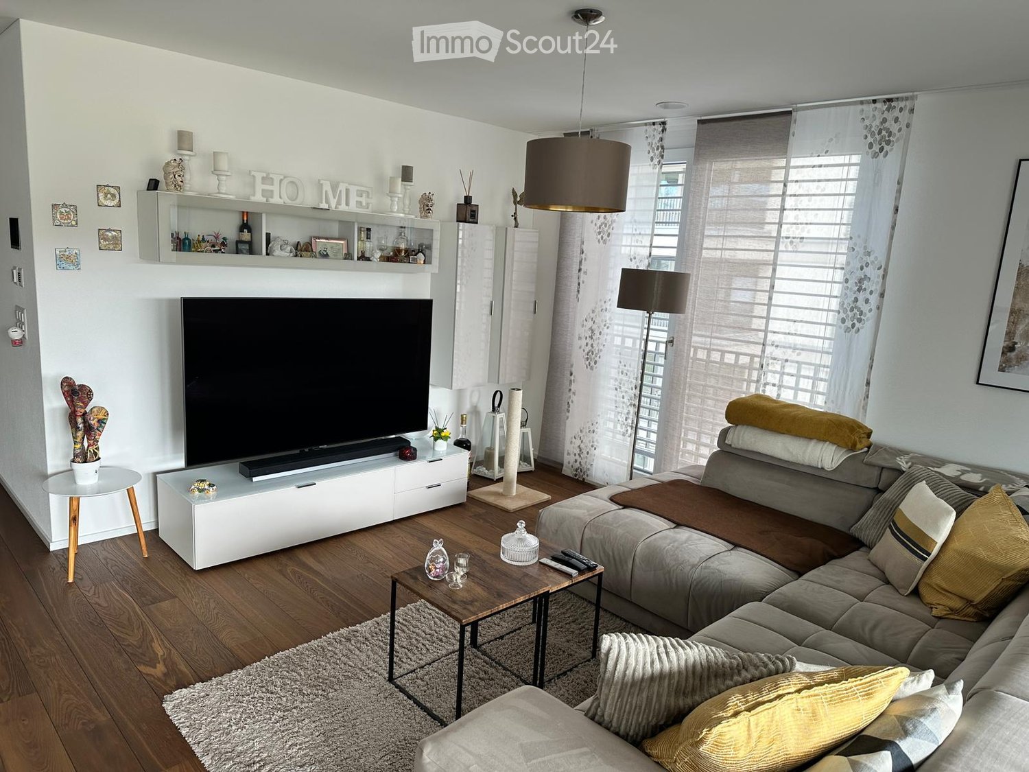 Modern living room with a grey sectional sofa, wooden coffee table, TV stand, cat tree, and decorative shelves. The room features large windows with sheer curtains and a pendant light hanging from the ceiling.