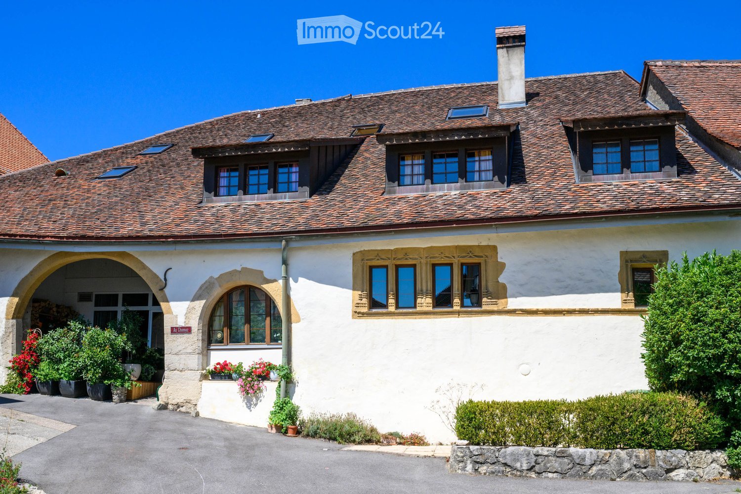 A two-story house with a tiled roof, white exterior walls, and multiple windows. The house features an arched entrance with potted plants and flowers, and is surrounded by well-maintained greenery and shrubs.