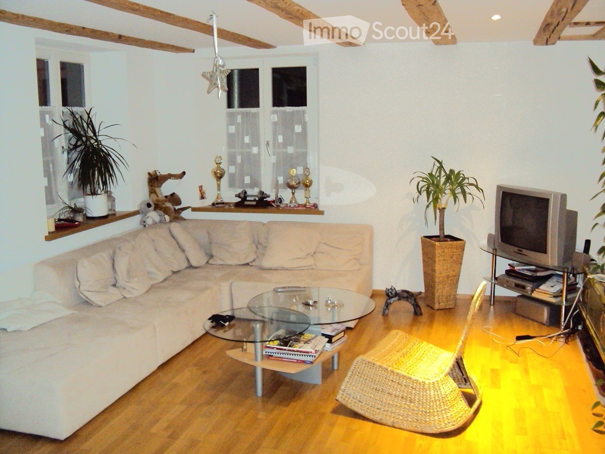 Living room with a white sectional sofa, glass coffee table, wicker chair, TV, and decorative plants on shelves.