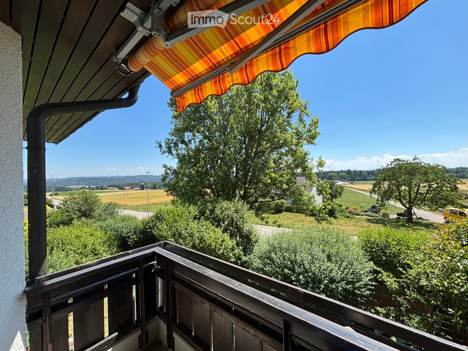 Balcony with wooden railing, striped awning, overlooking a rural landscape with fields, trees, and distant hills.