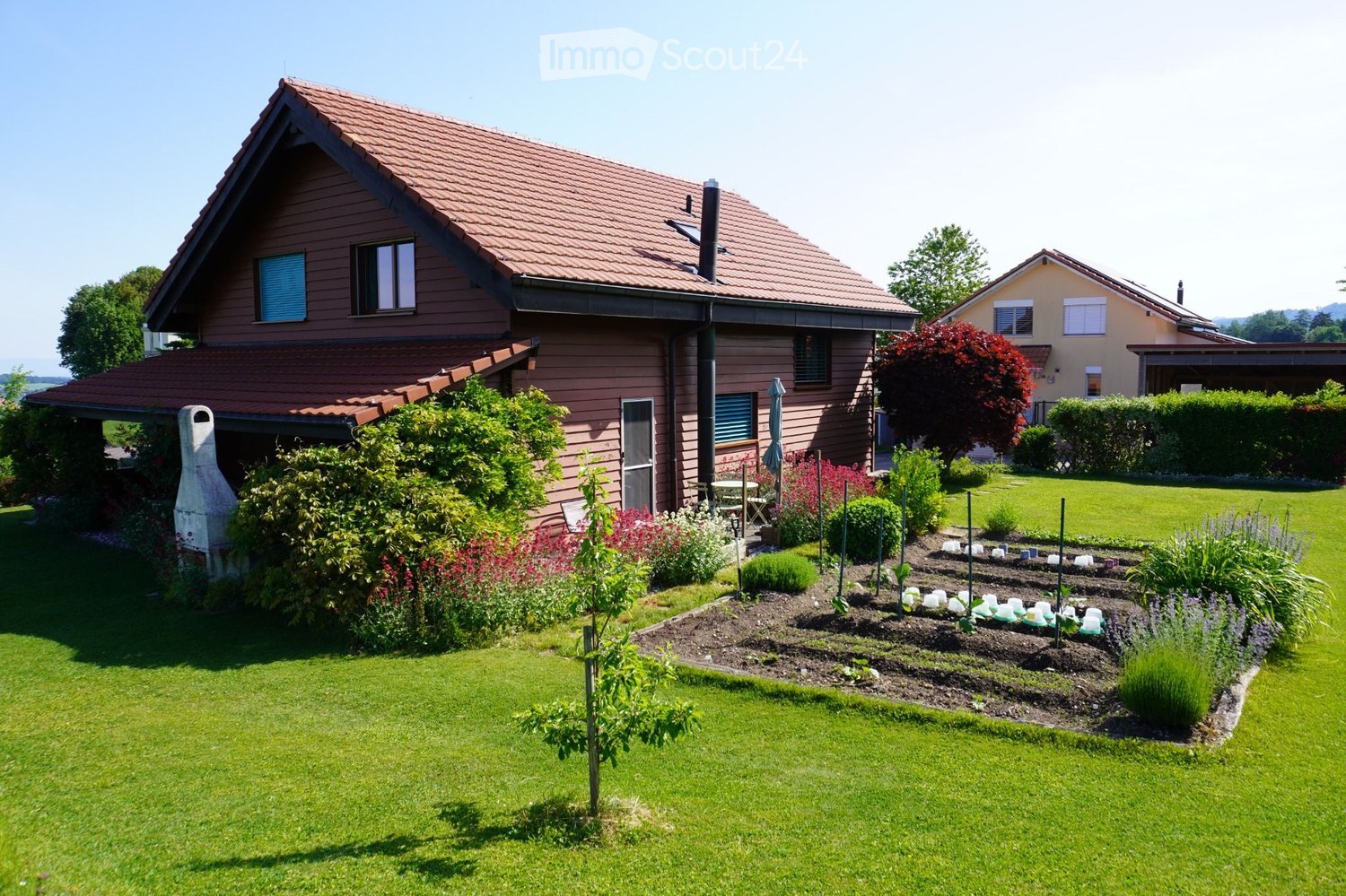 A well-maintained garden with a lawn, flower beds, a small tree, and a patio area with a table and chairs near the house.