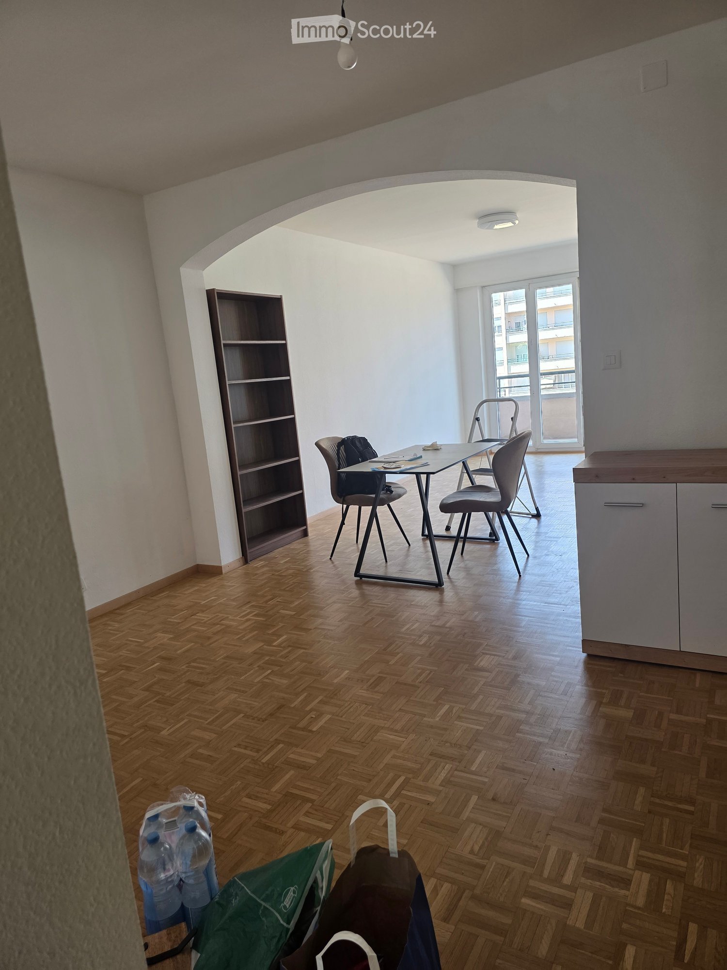 A modern, unfurnished room with wooden flooring, a table, chairs, and a shelf. The room has large windows and a balcony view.