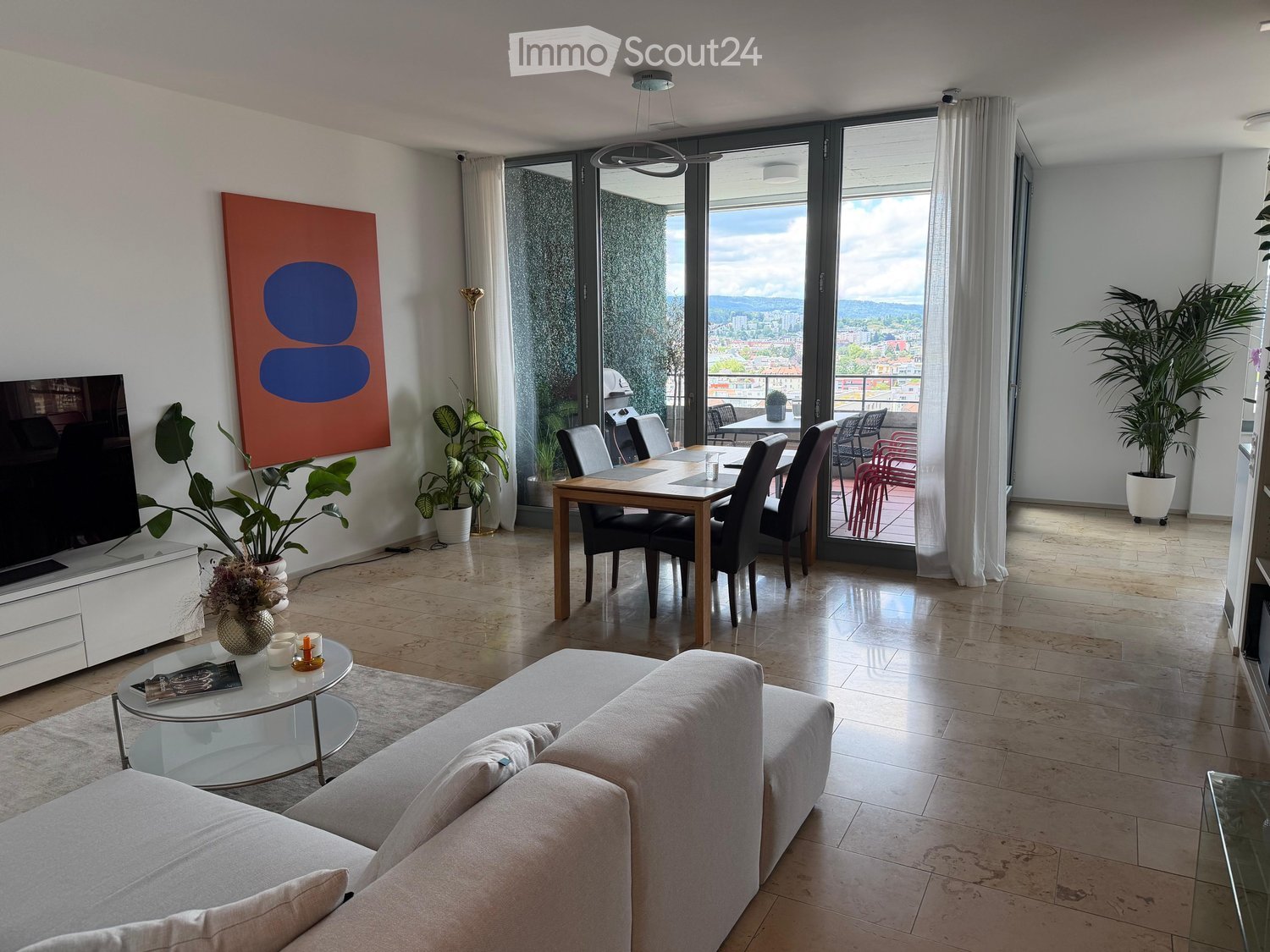 Modern living and dining area with a light grey sofa, glass coffee table, dining table with chairs, large windows with a view, and decorative plants.