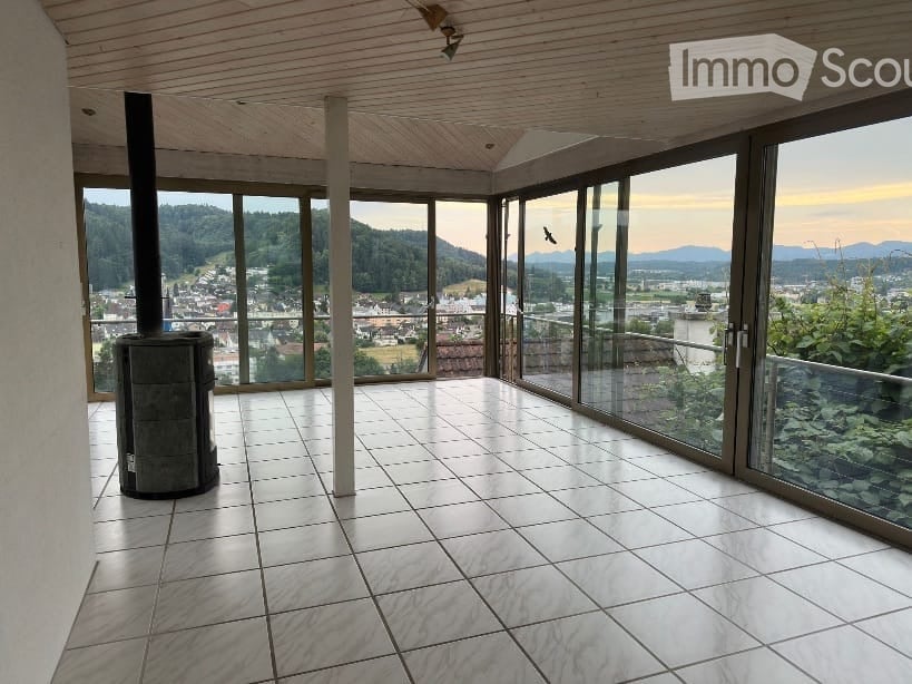 Bright, open space with large glass windows offering a scenic view of the valley and mountains. The room features a tiled floor and a wood-burning stove, with a wooden ceiling and modern lighting fixtures.