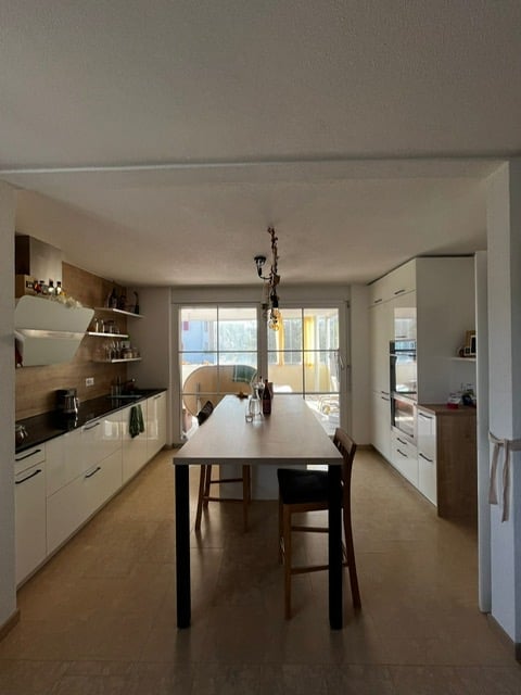 Modern kitchen with white cabinets, a dining table with chairs, and a view through large windows.