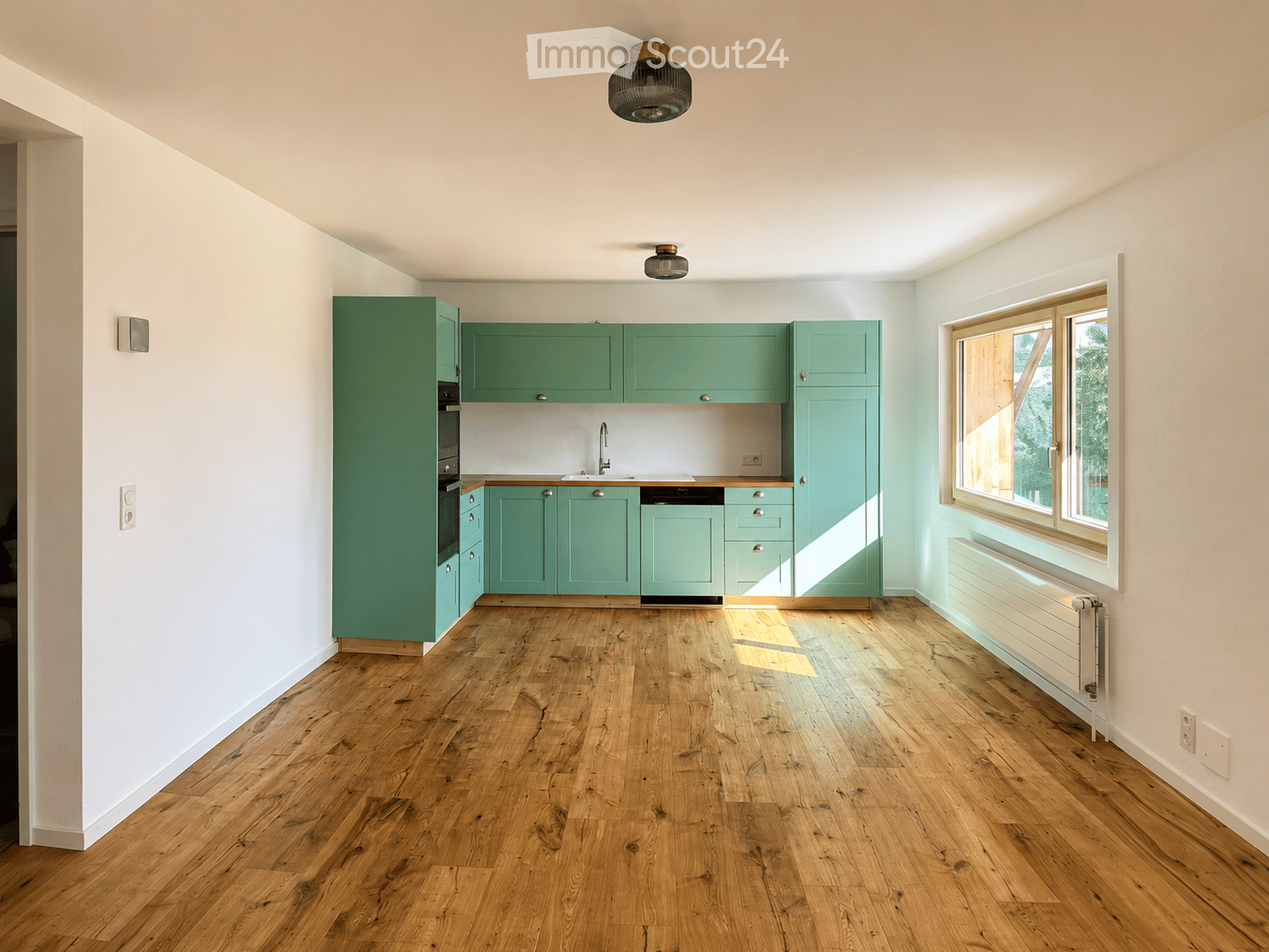 Empty kitchen with light green cabinets, wooden flooring, and a window letting in natural light.