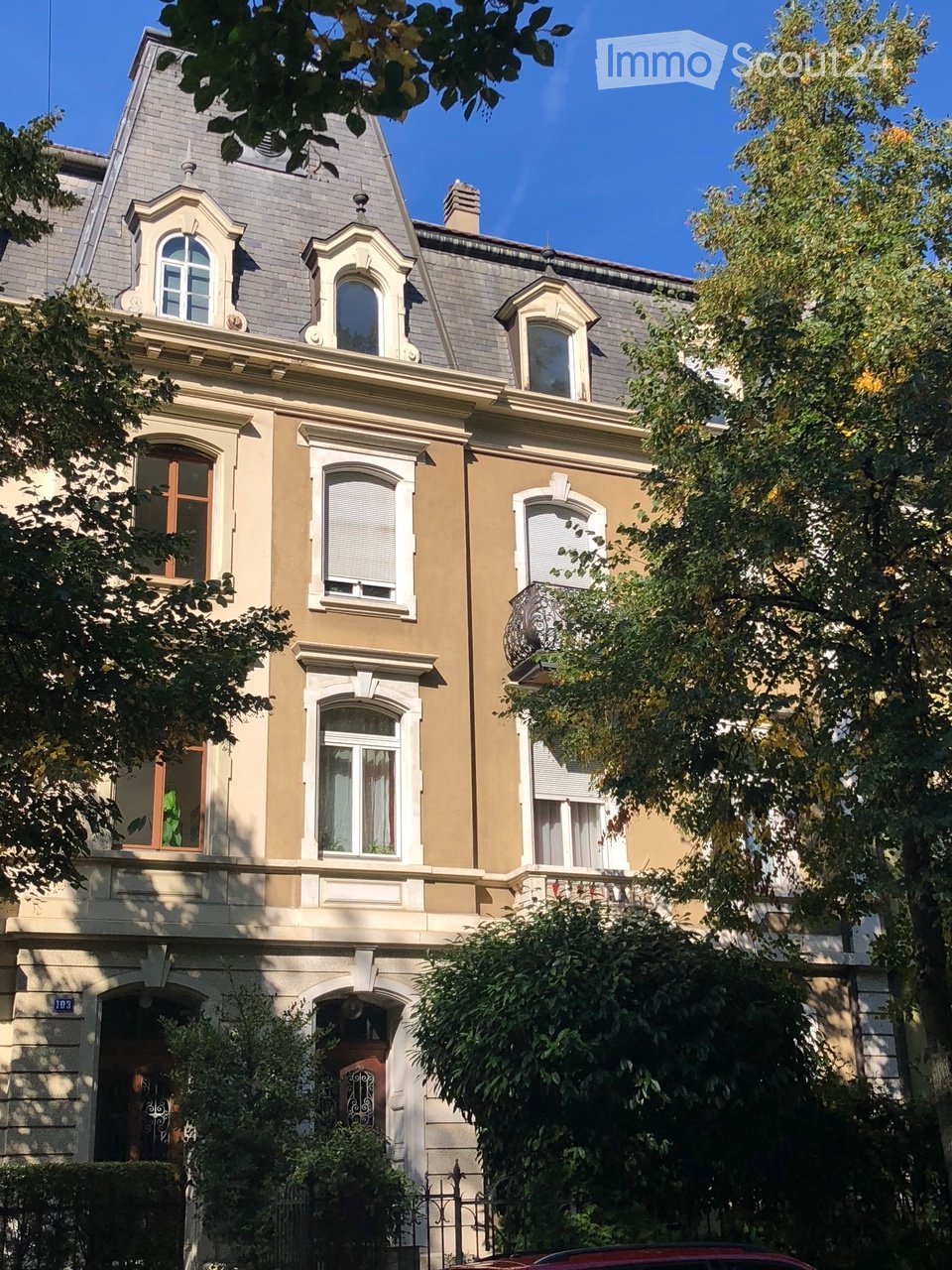 A multi-story residential building with beige and grey facade, arched windows, decorative moldings, and a grey roof with dormer windows.