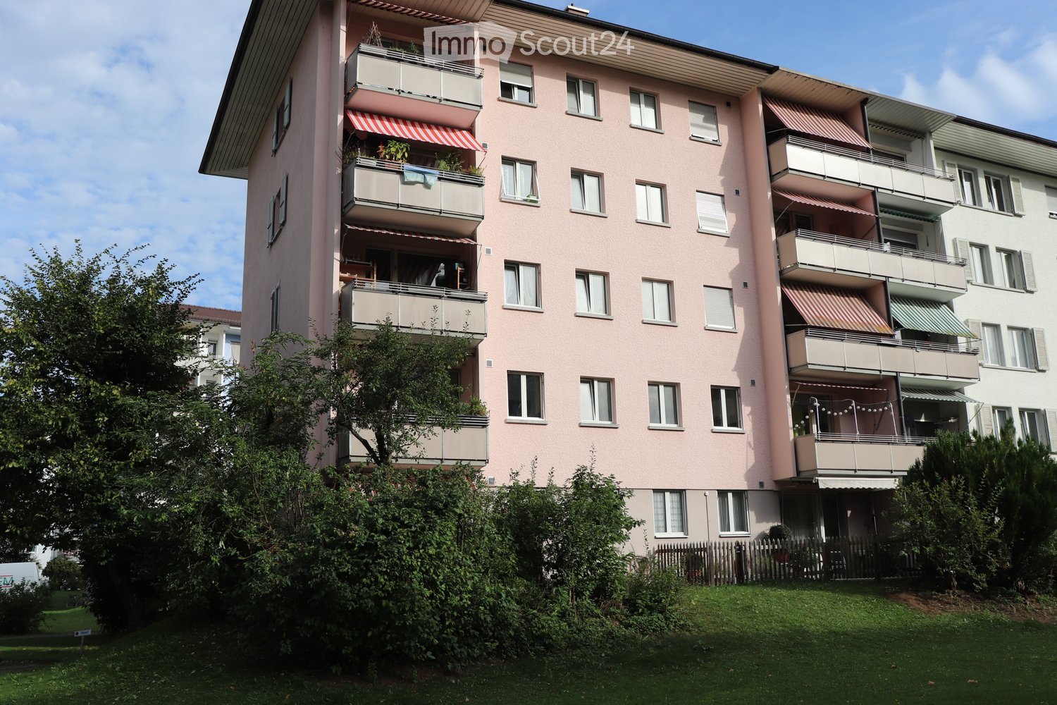 A multi-story residential building with pink facade, multiple balconies, and greenery in the foreground.