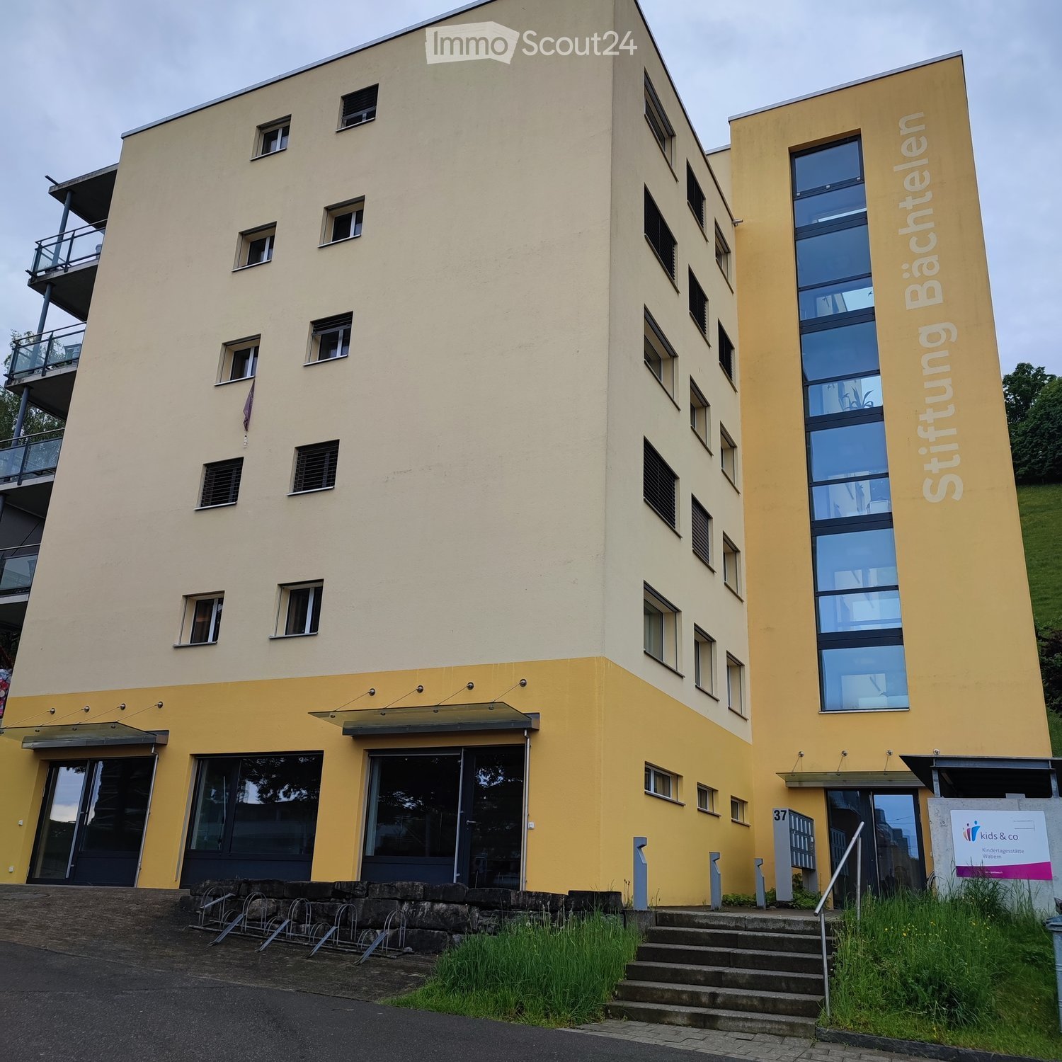 Modern multi-story building with yellow and beige facade, large windows, and a balcony on the upper floor.