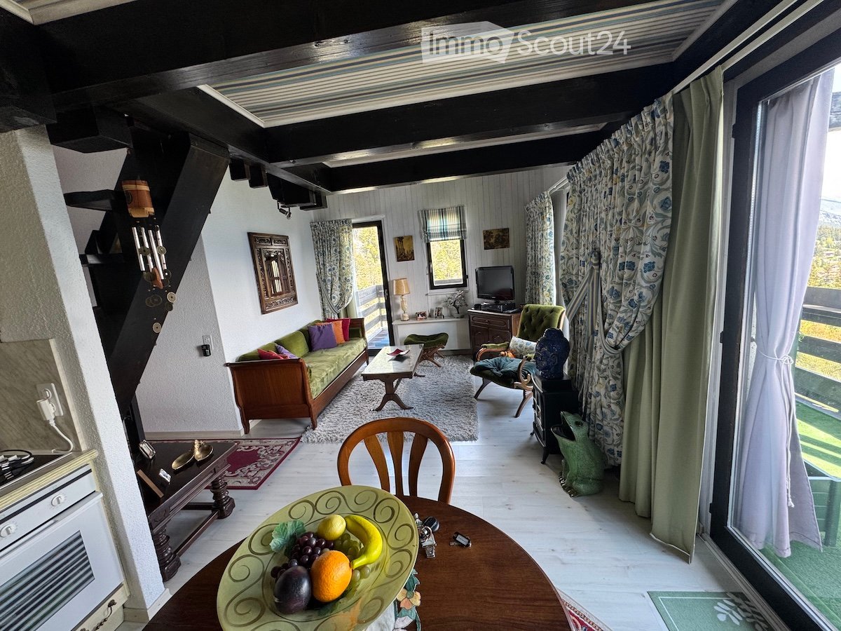 Bright living room with a green sofa, coffee table, wooden chairs, a TV, and a fruit bowl on a round table. Large windows offer a scenic view.