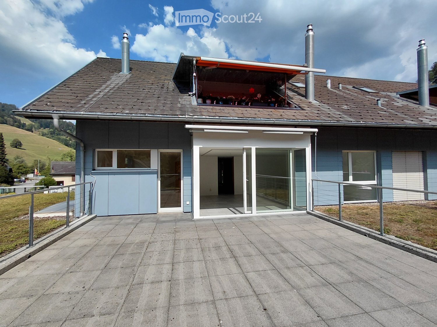 Two-story house with grey roof, light blue walls, large windows, and a covered balcony area with a railing.