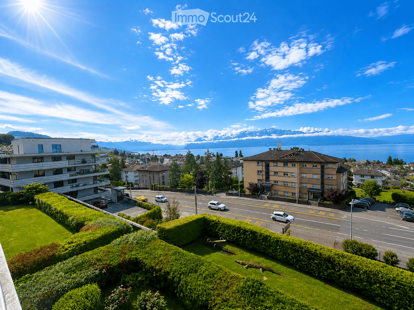 A panoramic view of a suburban area with multiple residential buildings, a road with cars, and a scenic view of a lake and mountains in the background.
