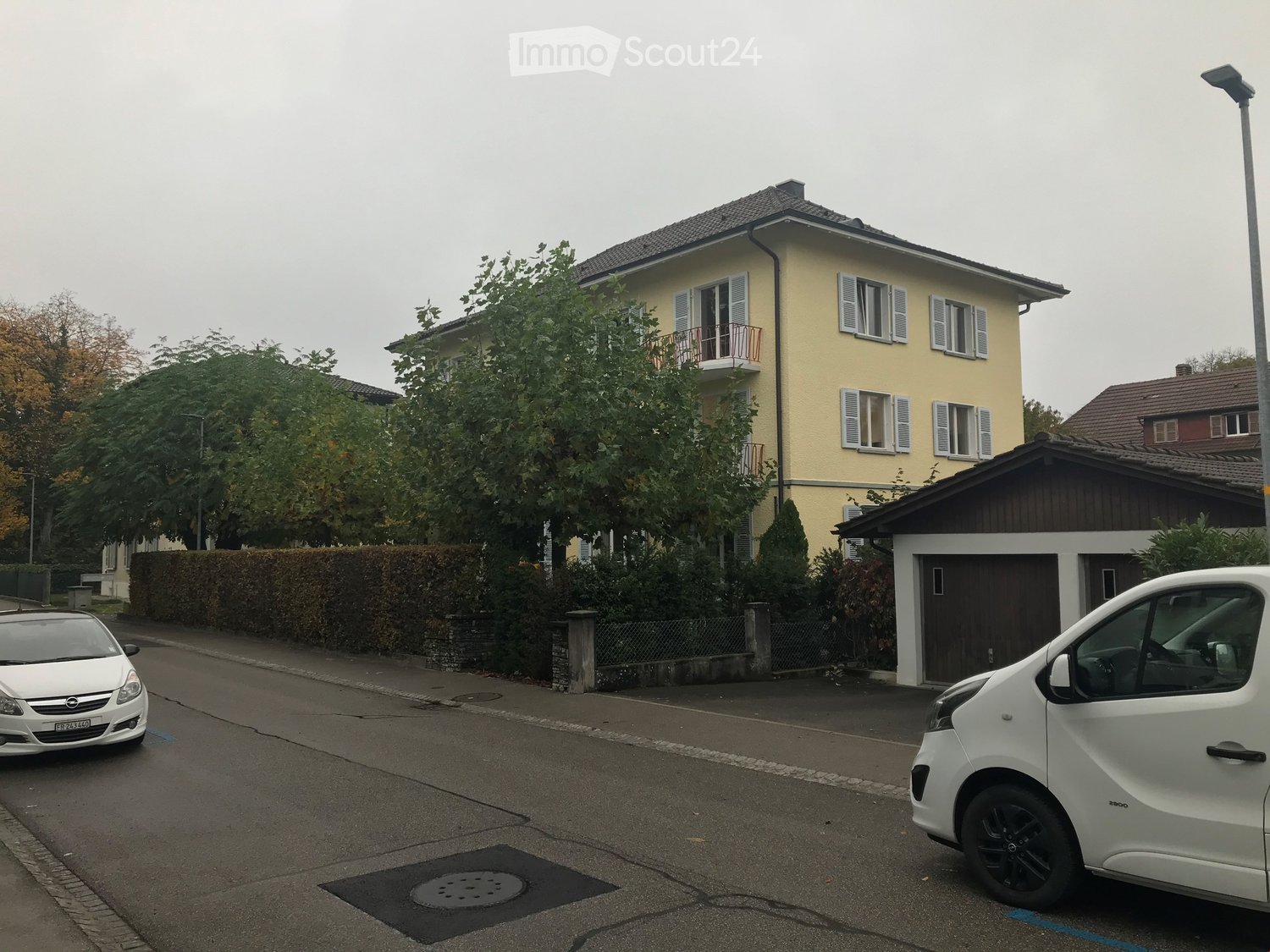 Two-story yellow house with white shutters, balcony, garage, and parked cars.