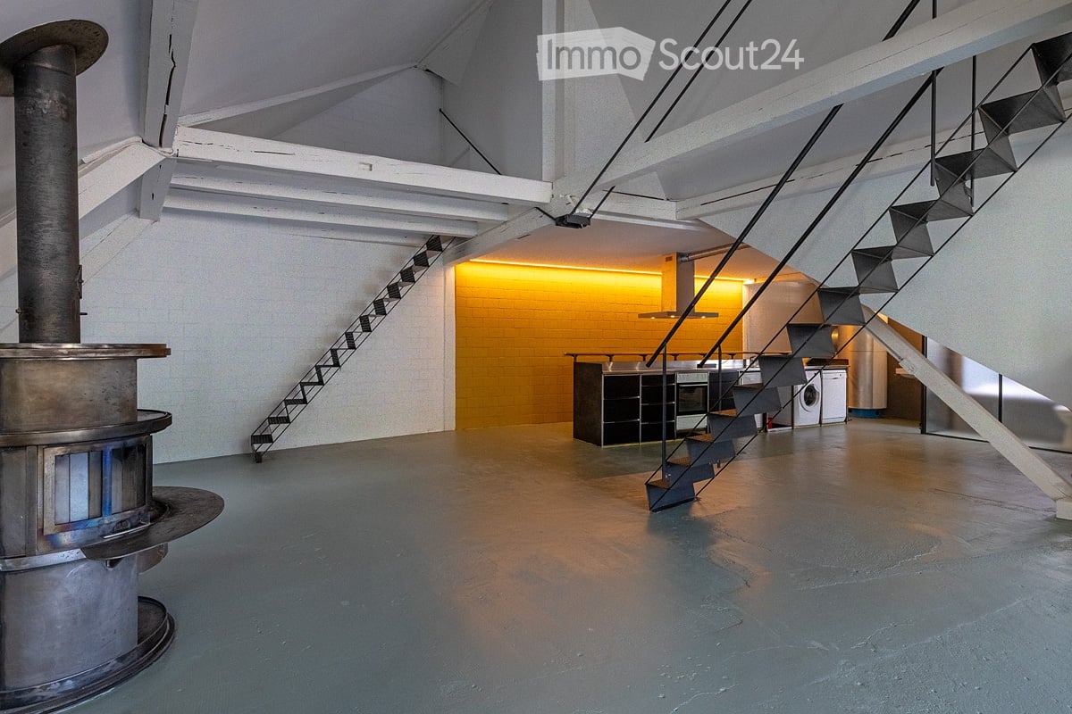 Open-plan space with a modern staircase, kitchen area featuring a central island, oven, and washing machine. Exposed metal chimney and industrial-style lighting.