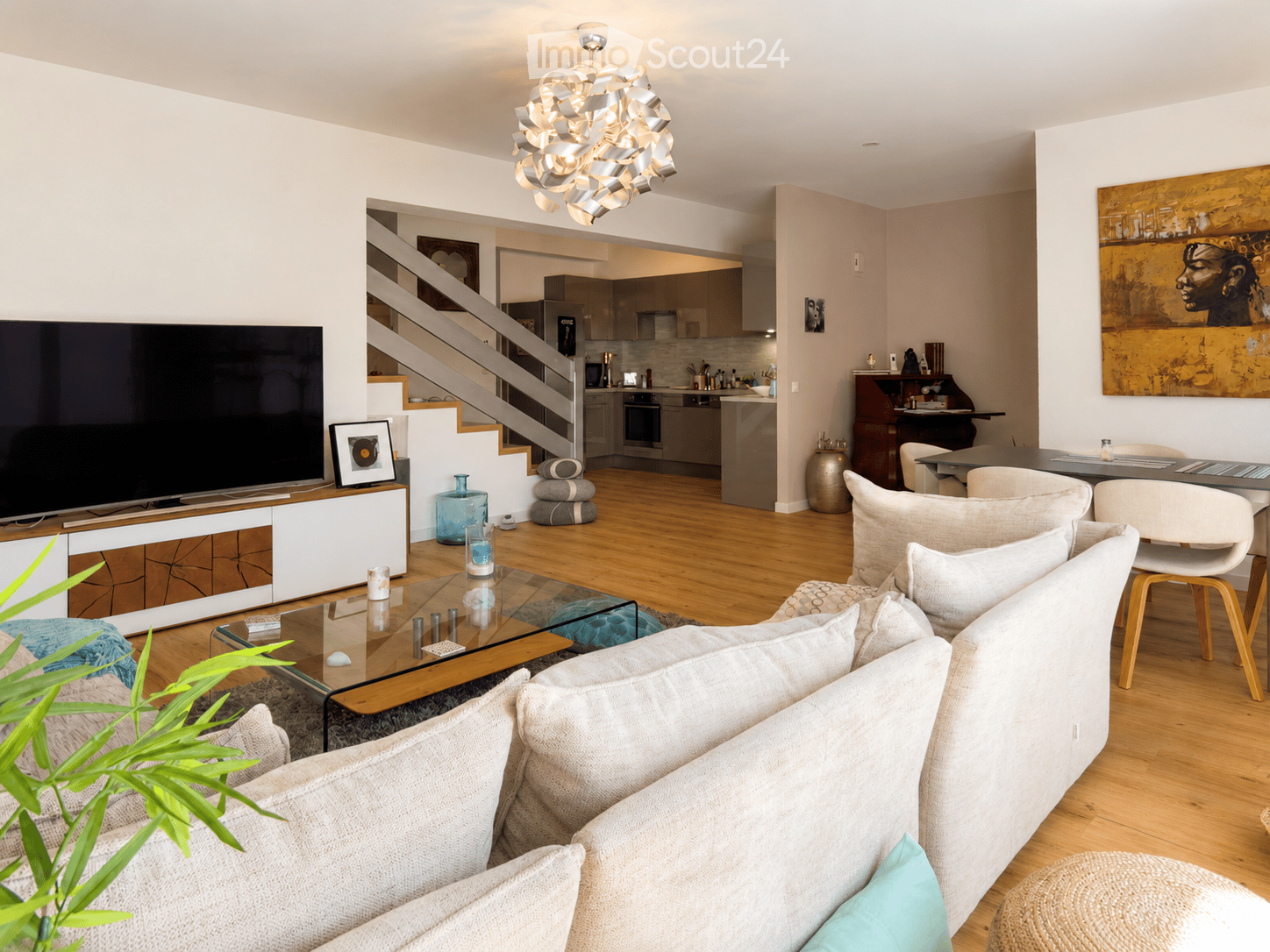 Modern living room with a white sofa, glass coffee table, and a TV stand. The room features a staircase leading to an open-plan kitchen area with modern appliances.