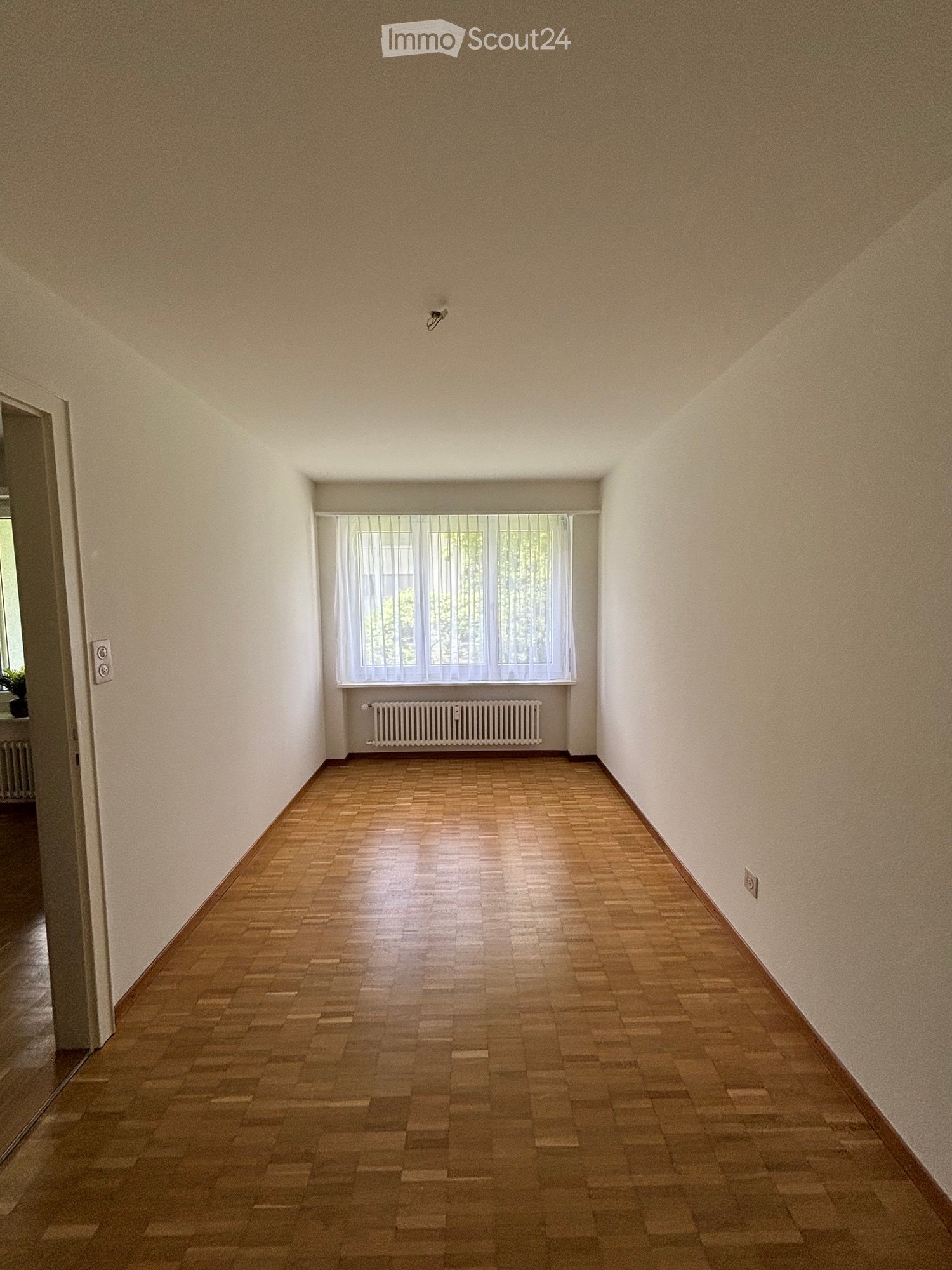 Empty hallway with wooden flooring, white walls, and a window with white curtains. A radiator is visible below the window.
